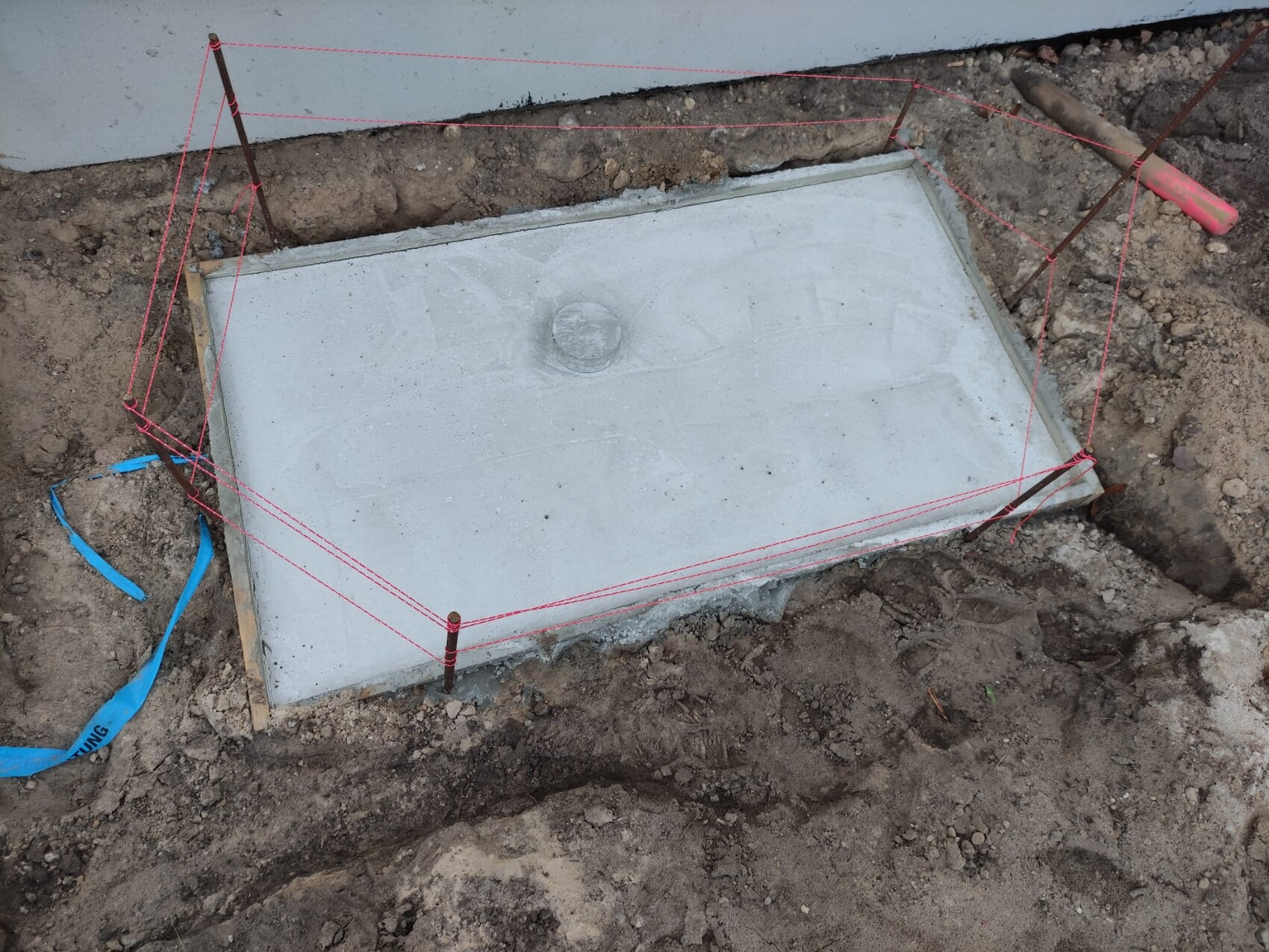 Rechteckige Betonplatte im Erdreich, von roten Messseilen umgeben auf einer Baustelle.
