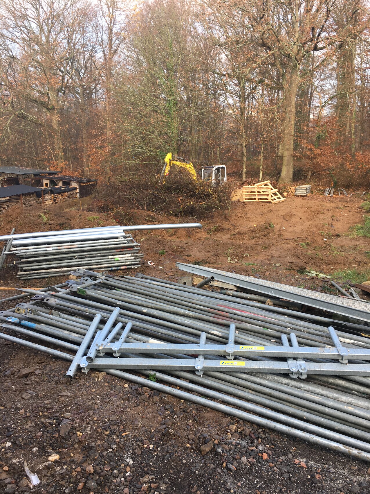 Baustelle im Wald: gestapelte Stahlrohre und Gerüste, gelber Bagger im Hintergrund, Paletten.