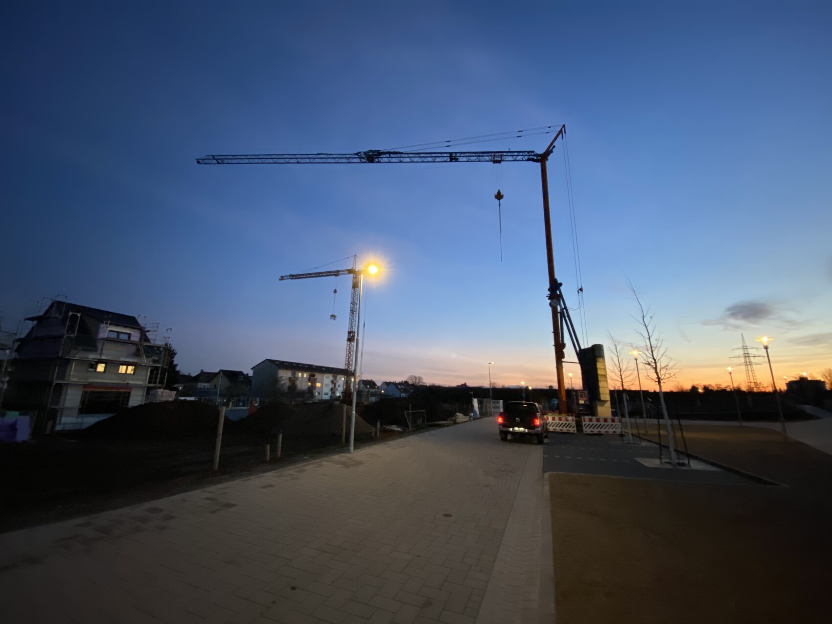 Abendliche Baustellenaufnahme: Krane, Fahrzeug, Bauzäune, Straßenlampen, Skyline im Dämmerlicht.
