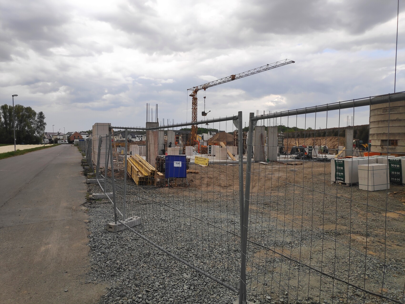 Baustelle mit Kran, Materialstapeln und Umzäunung; Straße links, bewölkter Himmel.