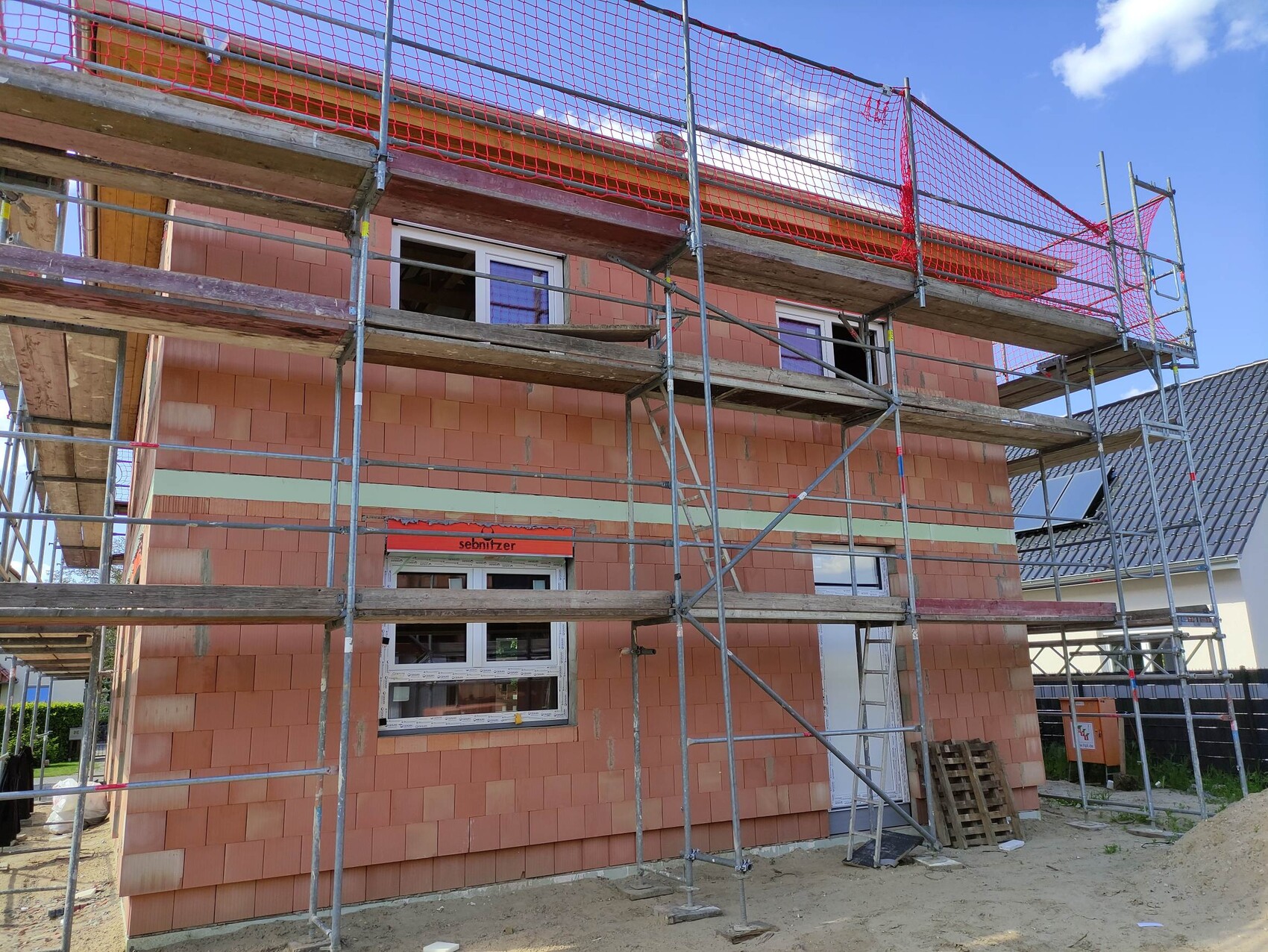 Neubau eines Hauses mit Gerüst, roten Sicherheitsnetzen, orange-braunen Ziegelwänden und offenen Fenstern.