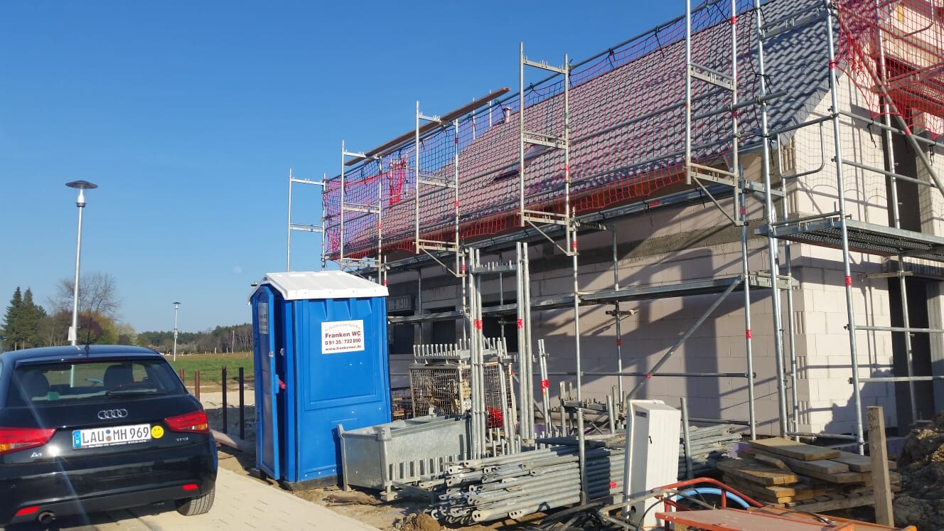 Baustelle mit Gerüst um ein Gebäude, blaue Bau-Toilette, Baumaterialien und geparktes Auto.