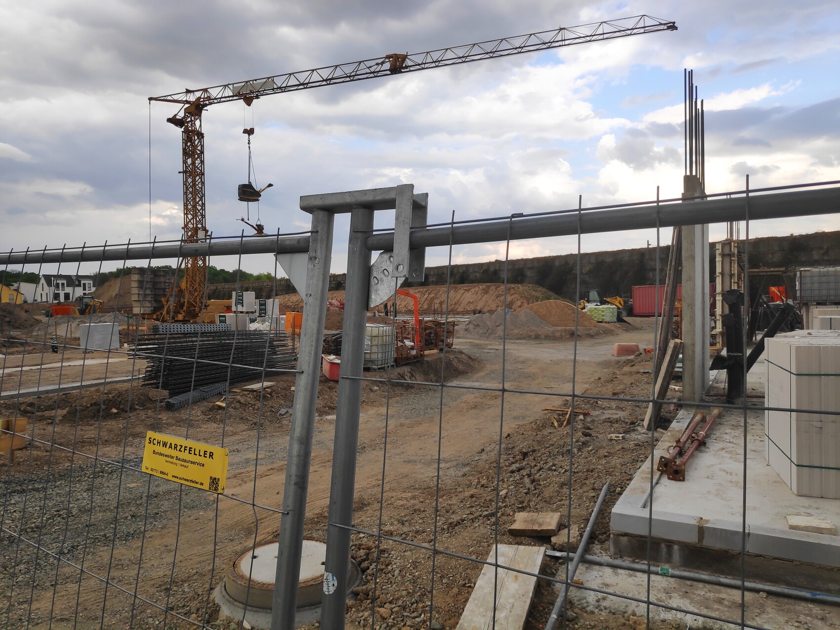 Baustelle mit Kran, Metallzaun und Materialstapeln im Vordergrund.