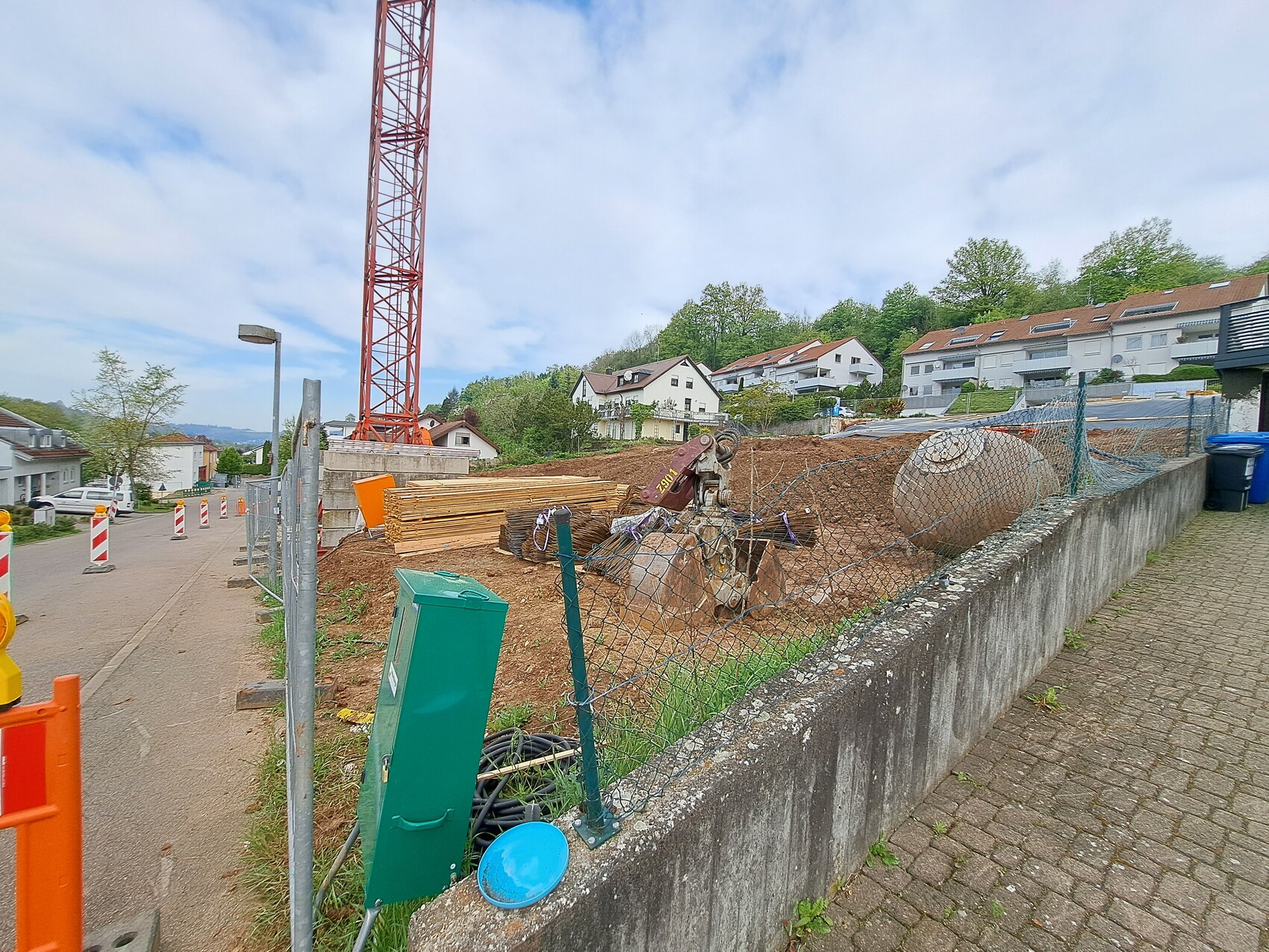 Baustelle mit rotem Kran, Holzstapeln, Betonmischer, Zaun und Wohnhäuser im Hintergrund