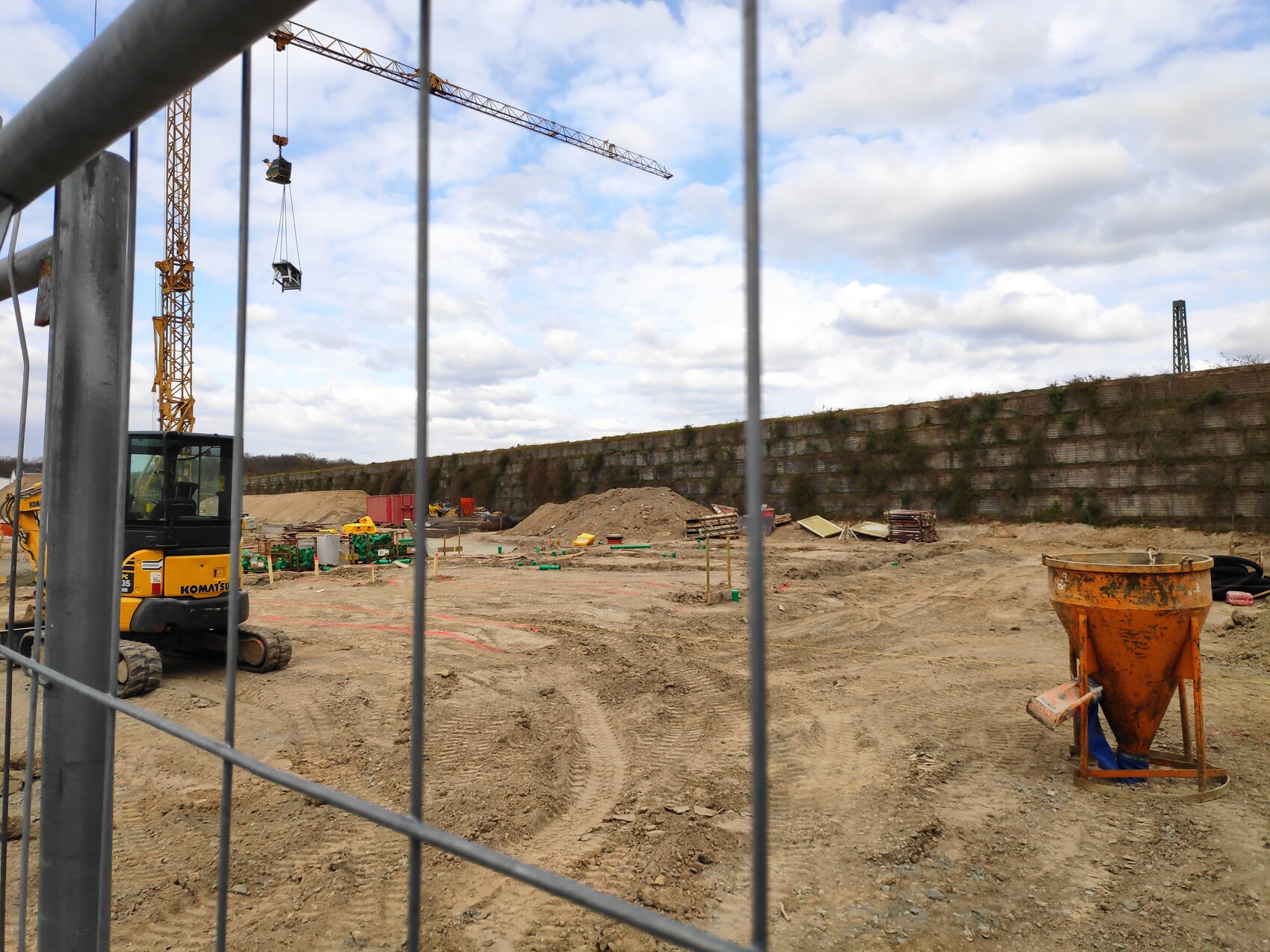 Baustelle mit Gelber Bagger, Turmdrehkran, Betonmischer und Sandhaufen vor Zaun.