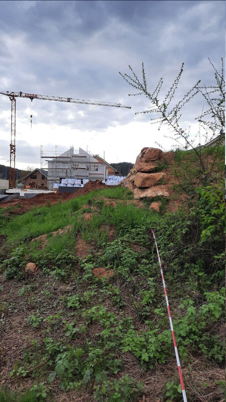 Kran über Baustelle mit Gerüst, Baufortschritt; grasiger Hang im Vordergrund