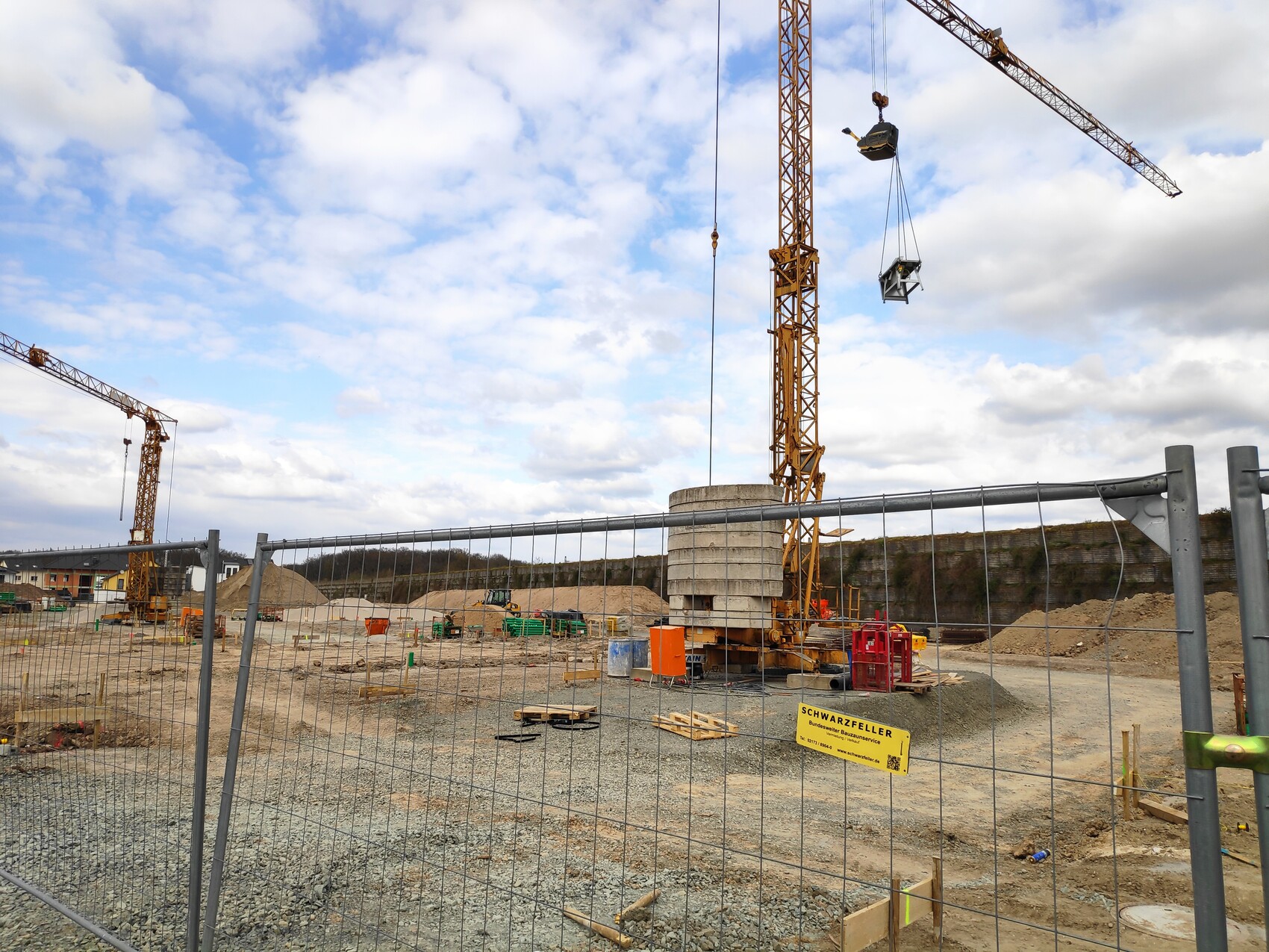 Große Baustelle mit zwei gelben Kränen, Baumaterialien und Zaun im Vordergrund.