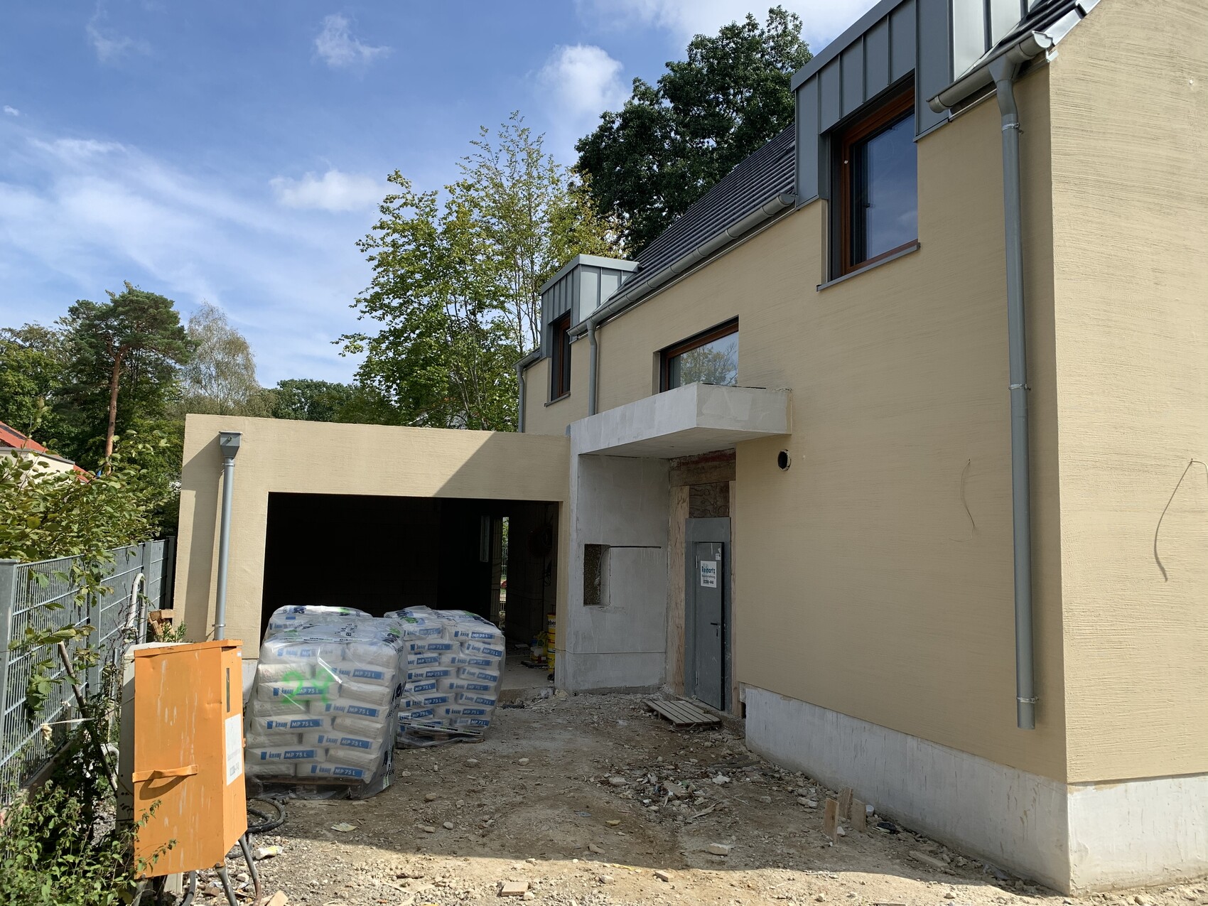 Baustelle vor beigeem Haus mit offener Garage, Paletten mit Zementsäcken und orangeem Container.