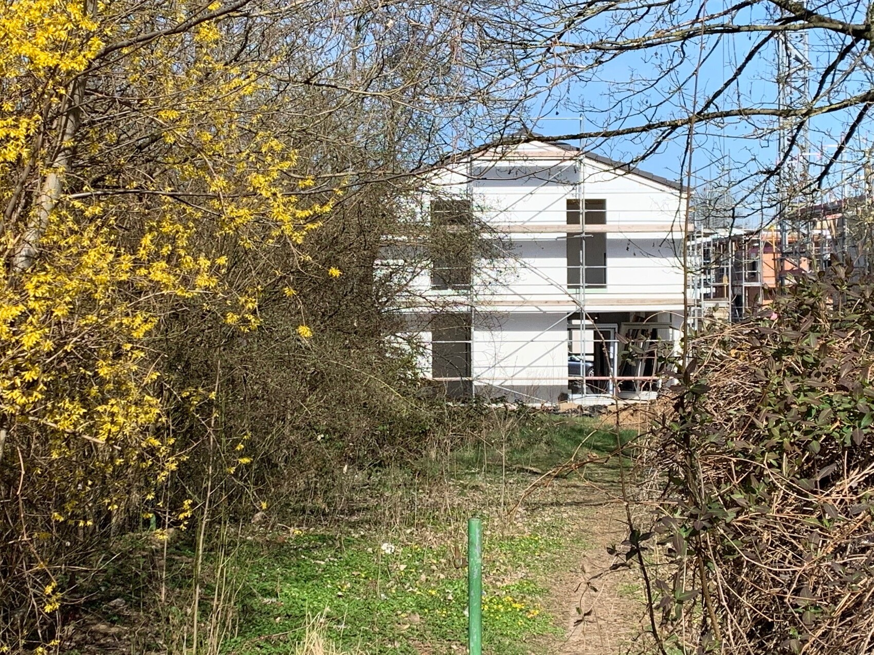 Weißes zweistöckiges Haus im Bau mit Gerüst, umliegendem Gelände und Bäumen im Vordergrund