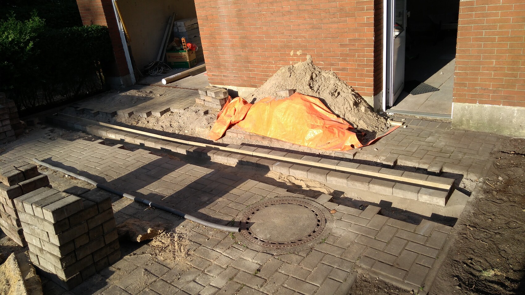 Baustelle vor Backsteinhaus: sandiger Haufen, orange Plane, neue Pflastersteine, offener Eingang