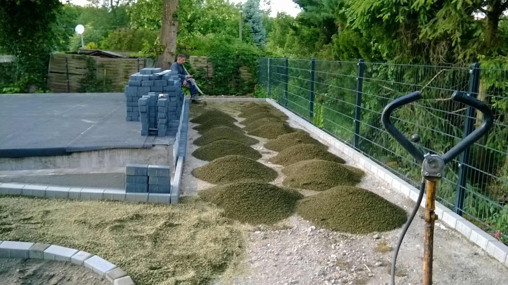 Mann sitzt neben gestapelten Betonpflastersteinen; Kieshaufen entlang eines Weges, Zaun rechts.