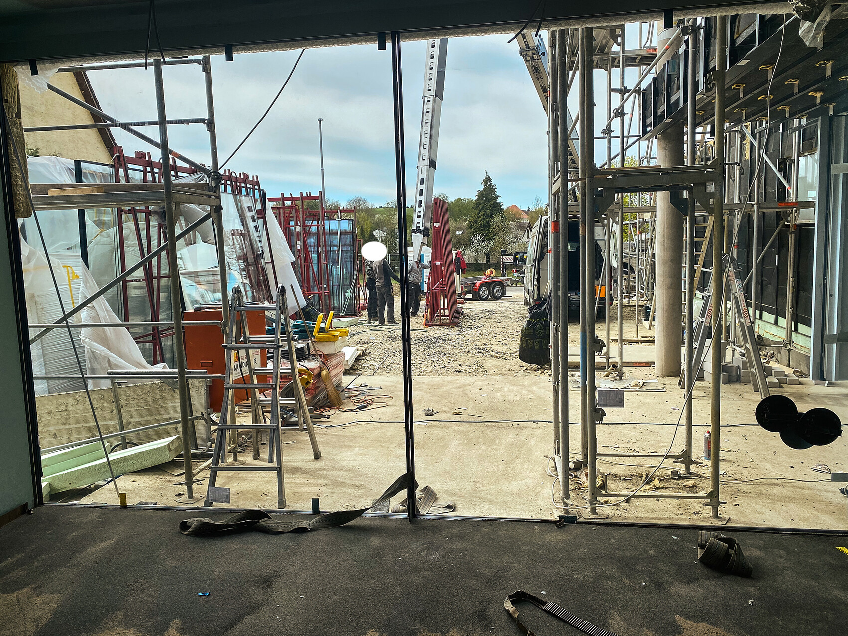 Blick durch ein offenes Baugerüst auf eine Baustelle mit Gerüsten, Geräten und Fahrzeugen.