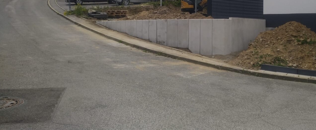 Straße vor Baugelände mit Betonstützmauer, Erdhügel und Bagger im Hintergrund.