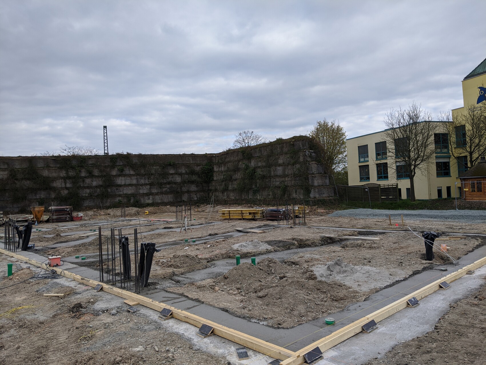 Baustelle: Fundamentgräben, Bewehrung; Felswand, Bäume, gelbes Gebäude im Hintergrund.