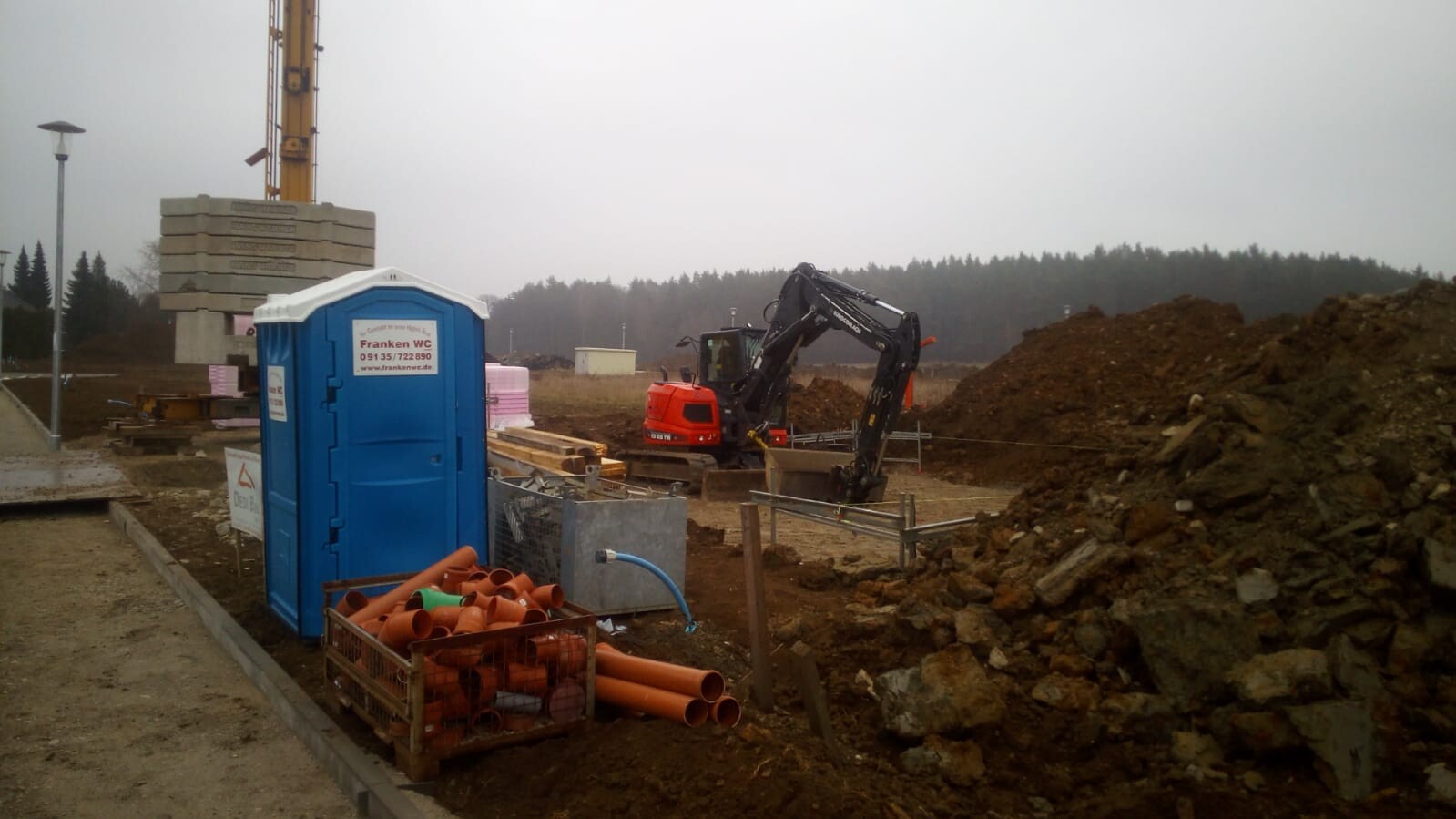 Bauplatz mit blauer Bau-WC-Kabine, Bagger, Rohrleitungen und Erdhaufen.
