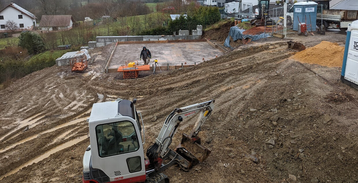 Baustelle an Hang mit Minibagger, Fundamentplatte wird vorbereitet, Arbeiter und Materialstapel sichtbar.