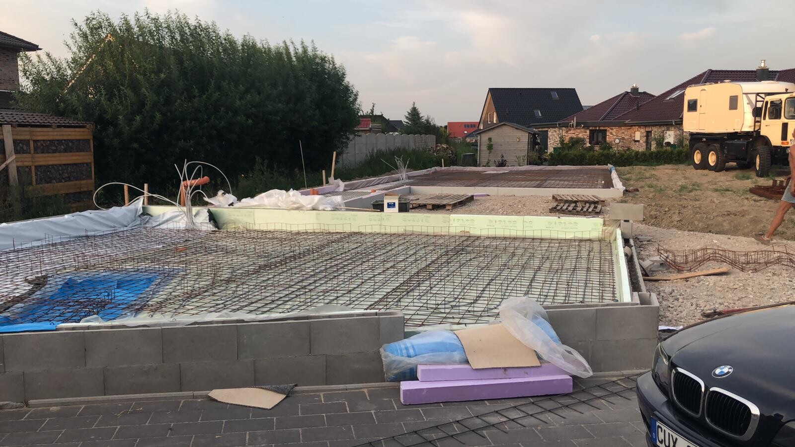 Fundament mit Stahlbewehrung auf Wohnhaus-Baustelle, sichtbare Baugeräte und Wagen im Hintergrund.