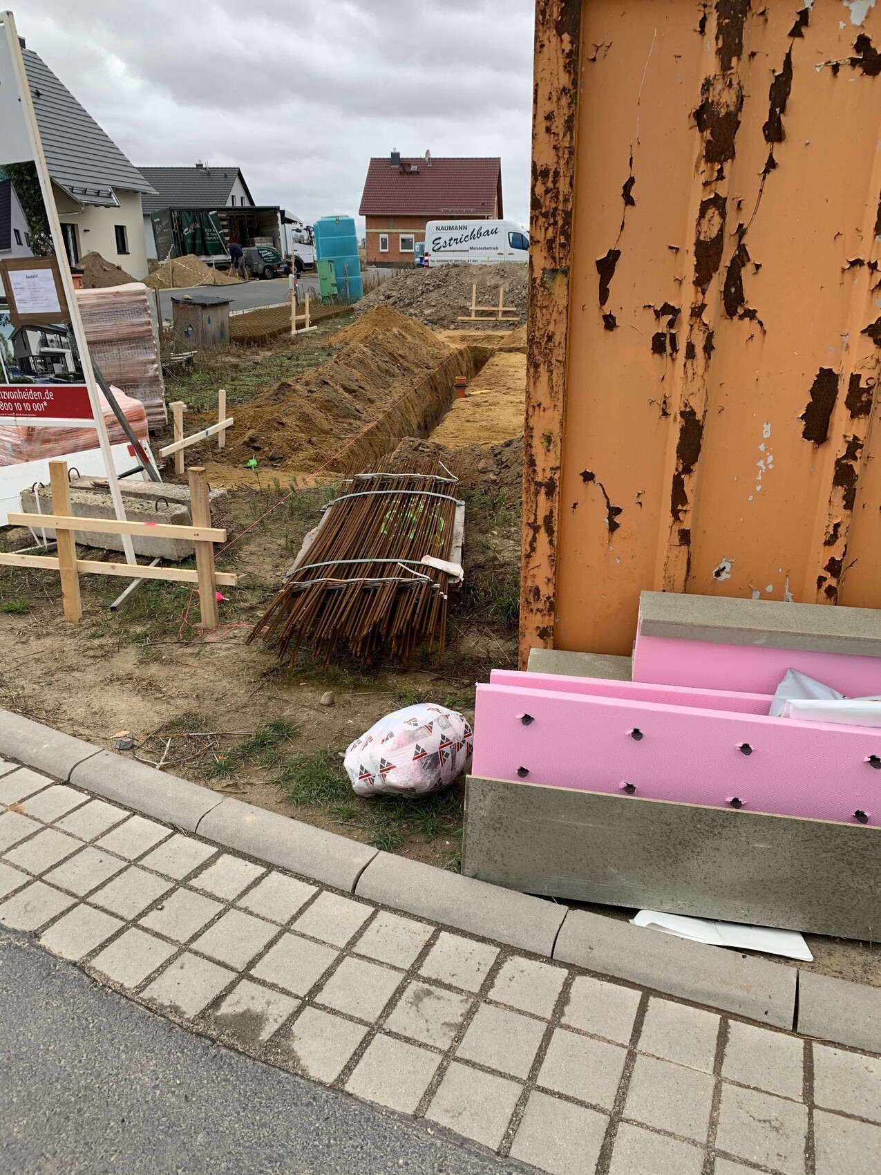 Baustelle in Wohngebiet: Bewehrungsstahl, rosa Dämmplatten, rostiger Pfeiler, Wagen im Hintergrund.
