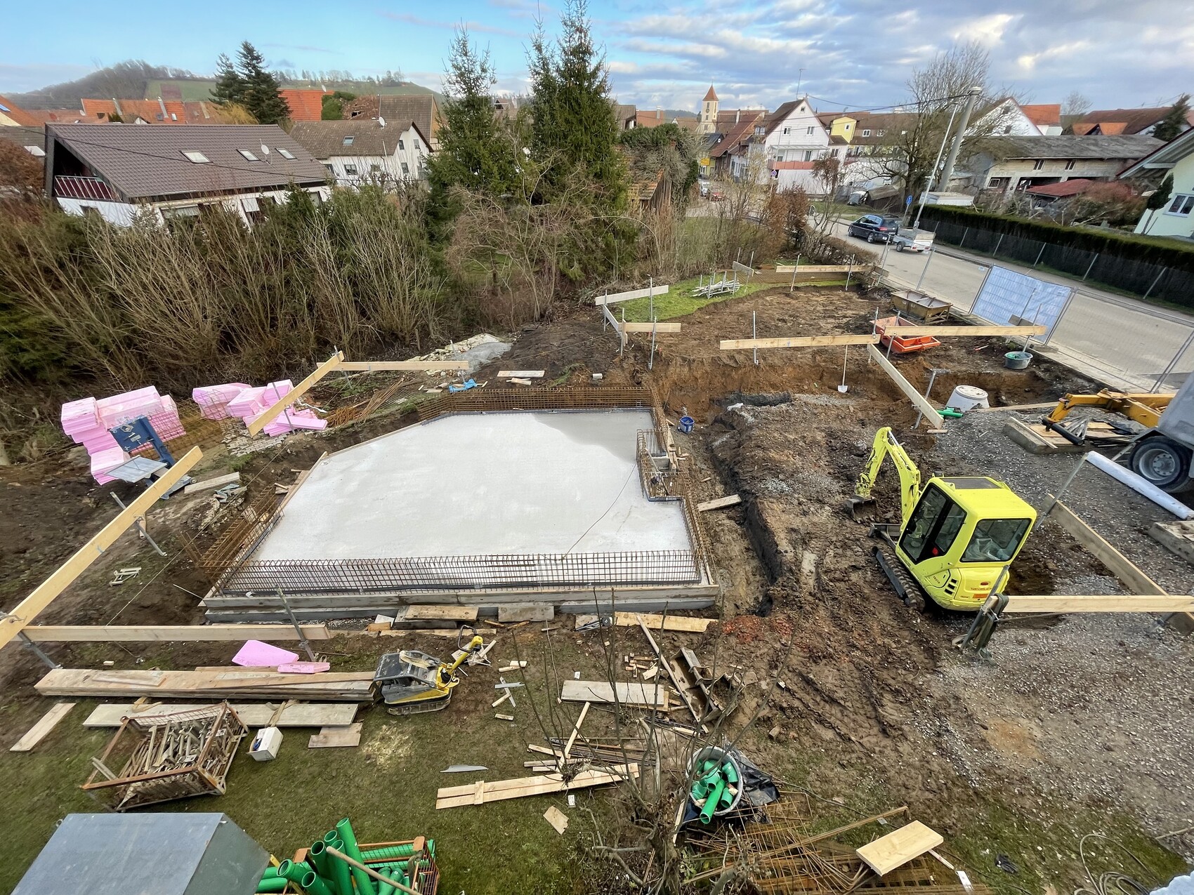 Baustelle im Wohngebiet: Betonbodenplatte mit Holzformen, Mini-Bagger und Baumaterialien.