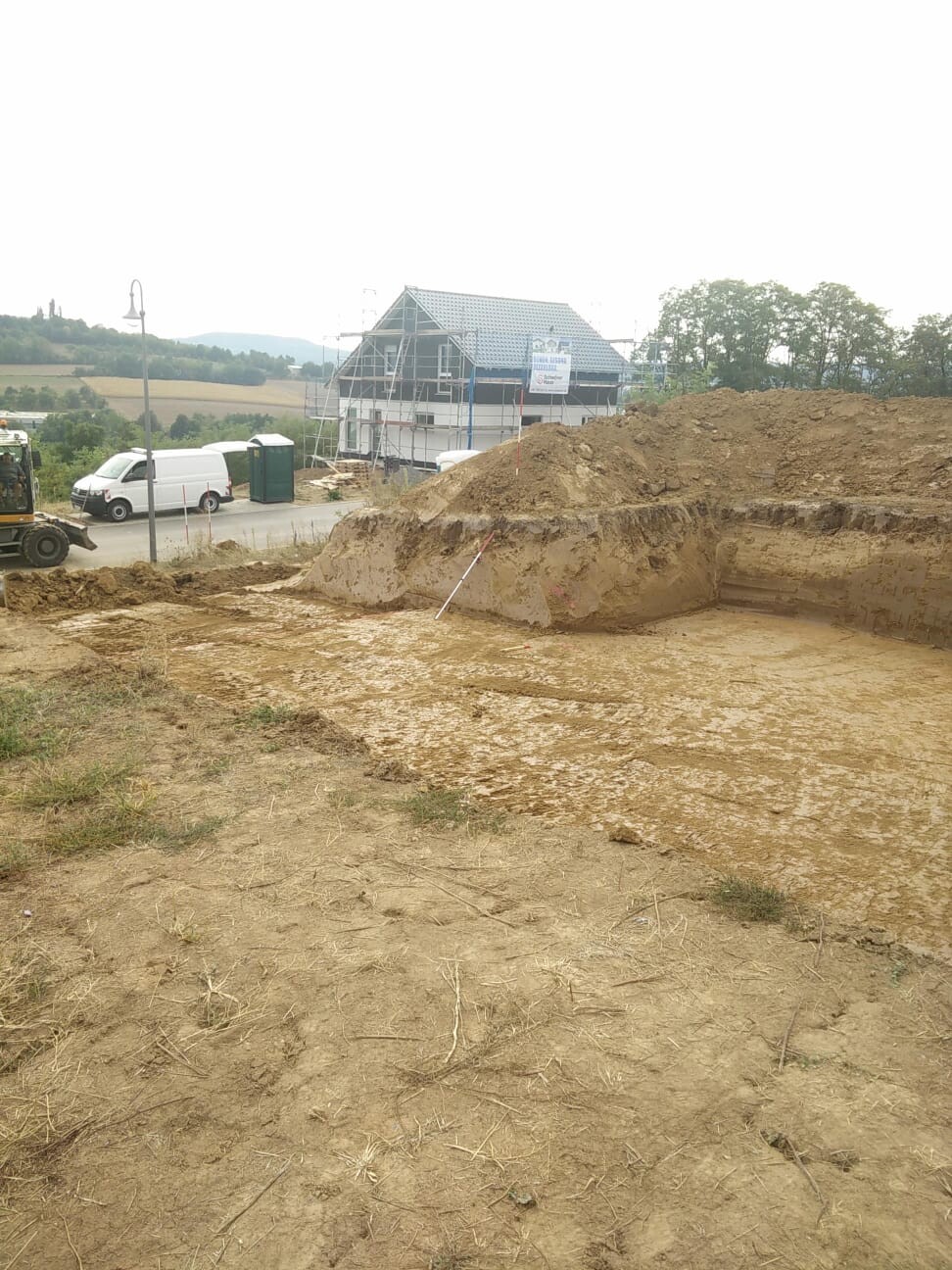 Baustelle mit Rohbau, großer Erdaufschüttung und Haus im Hintergrund