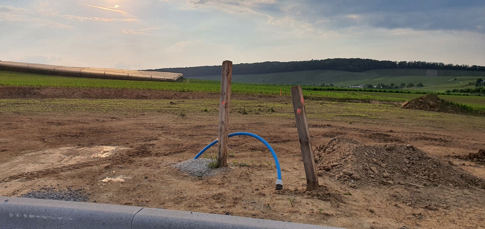 Ländliche Baufläche mit zwei Holzpfosten, blauem Schlauchbogen, Erde und Feldern im Hintergrund.