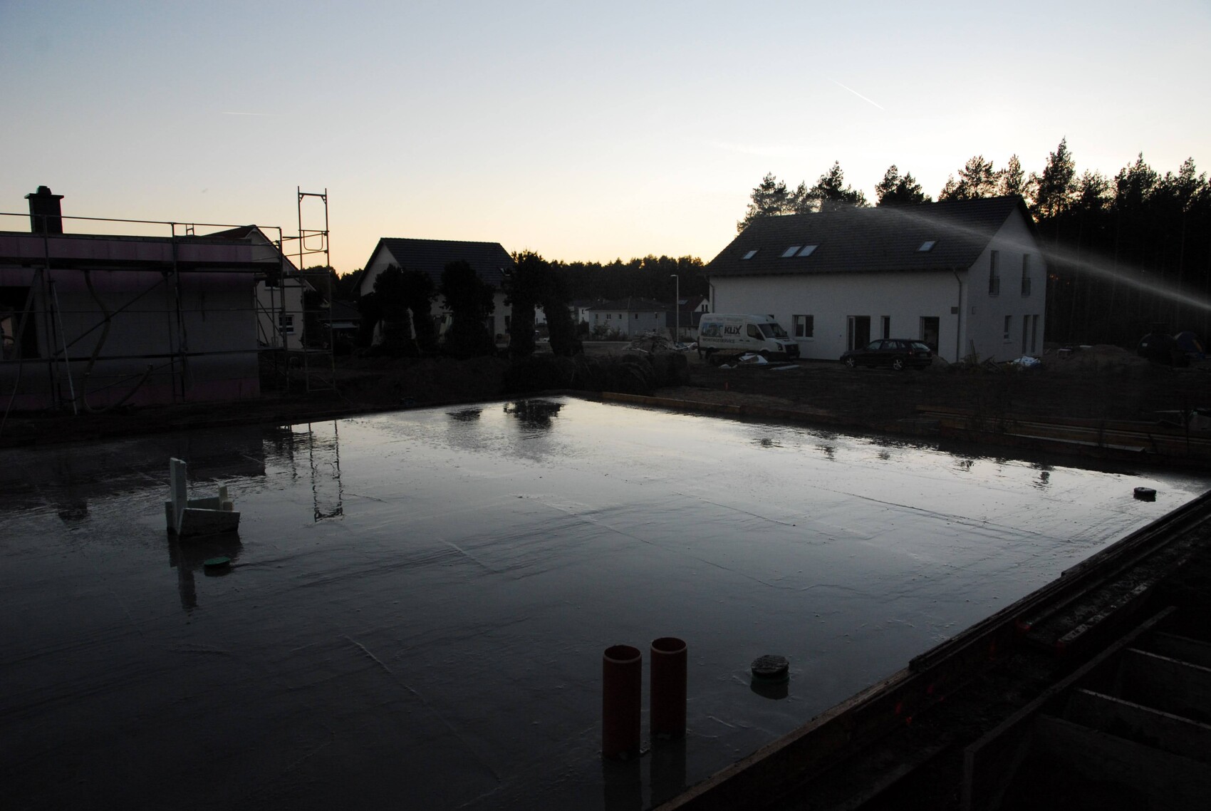 Baustelle mit flacher Wasseroberfläche, links Gerüst, rechts Wohnhäuser und Wald im Hintergrund.