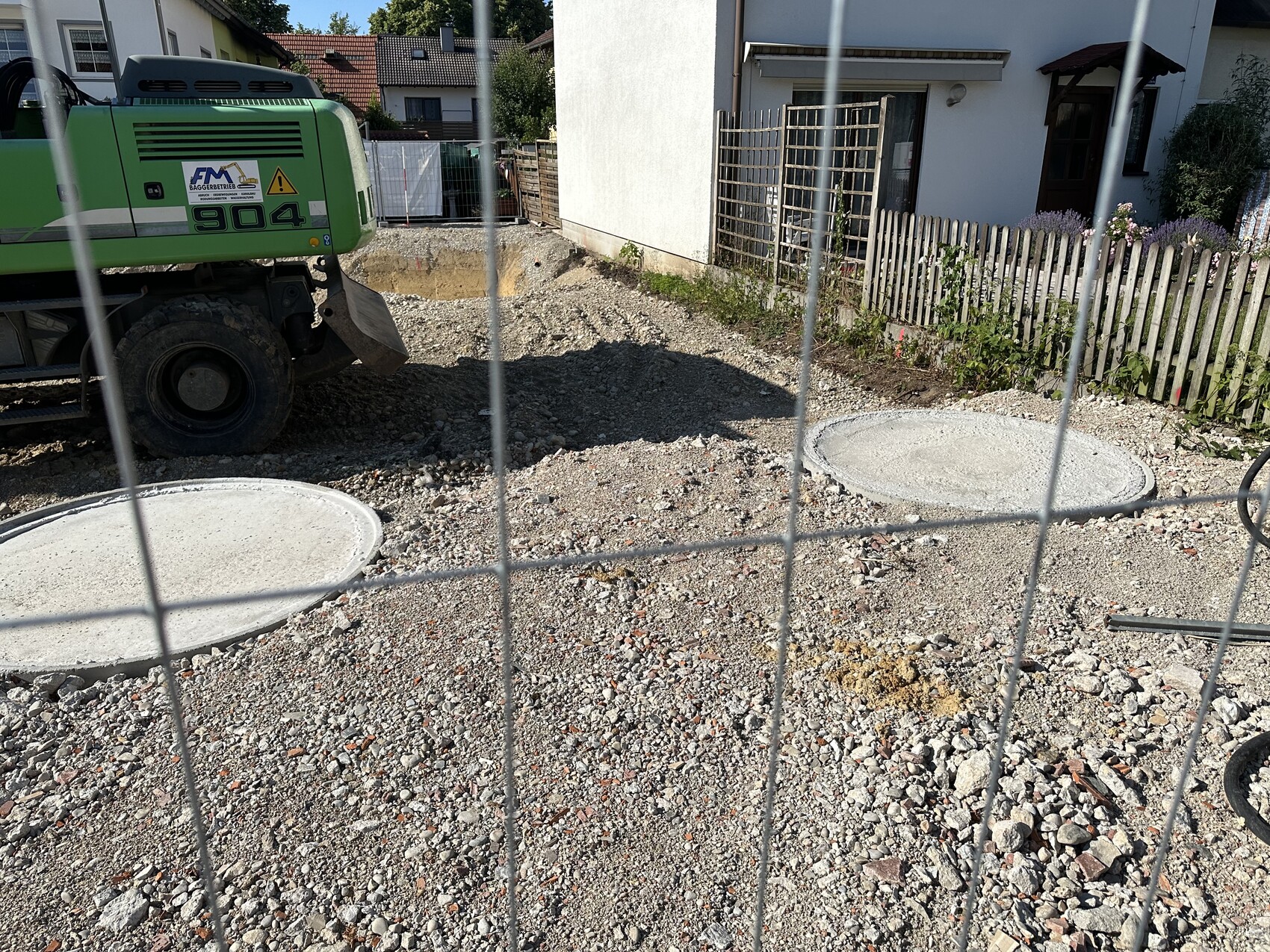 Baustelle vor Wohnhaus: grüne Baumaschine, offene Grabung, zwei runde Betondeckel, Gartenzaun.