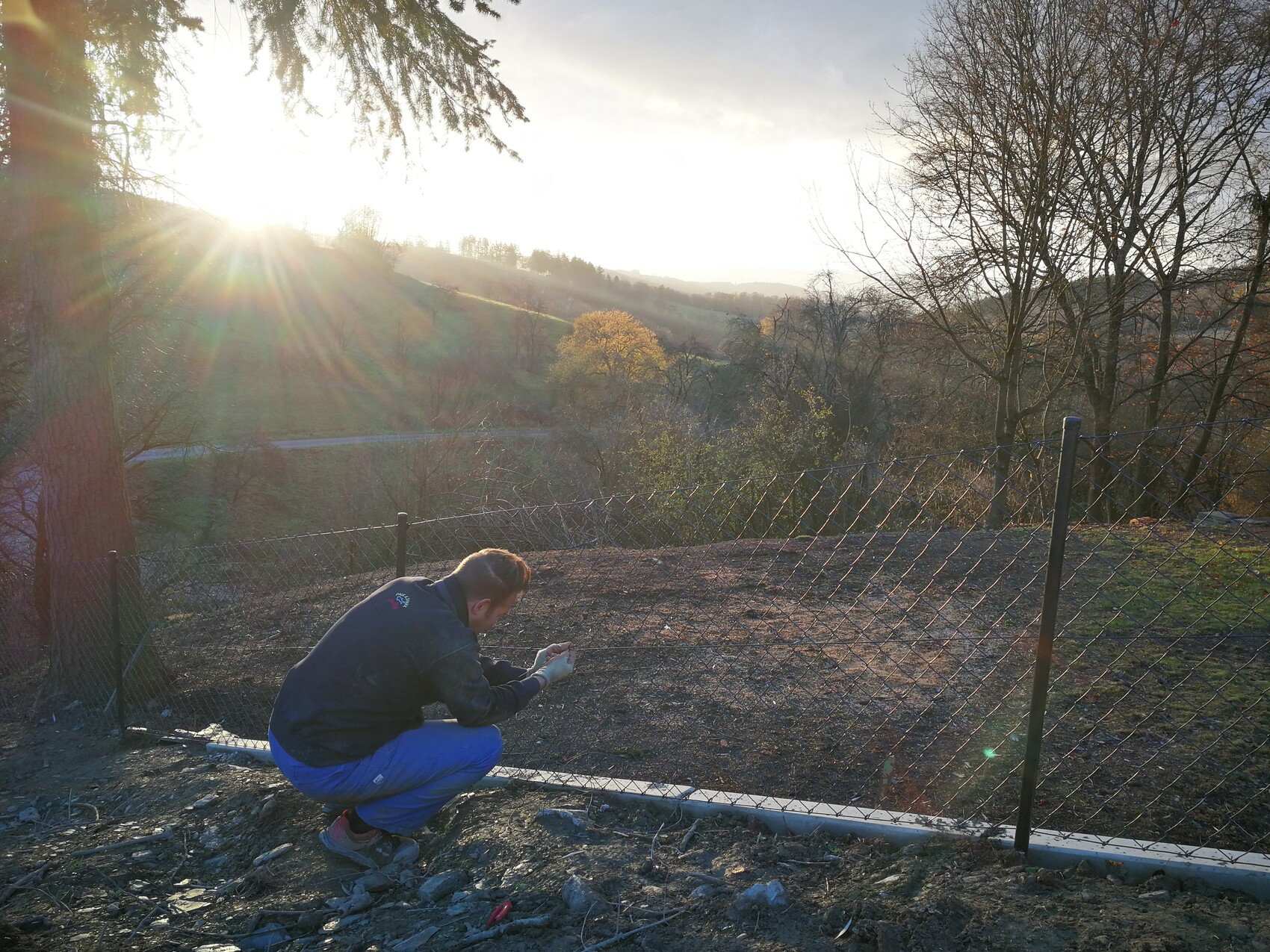 Mann kniet neben einem Maschendrahtzaun und arbeitet am Zaun in einer sonnigen Herbstlandschaft.