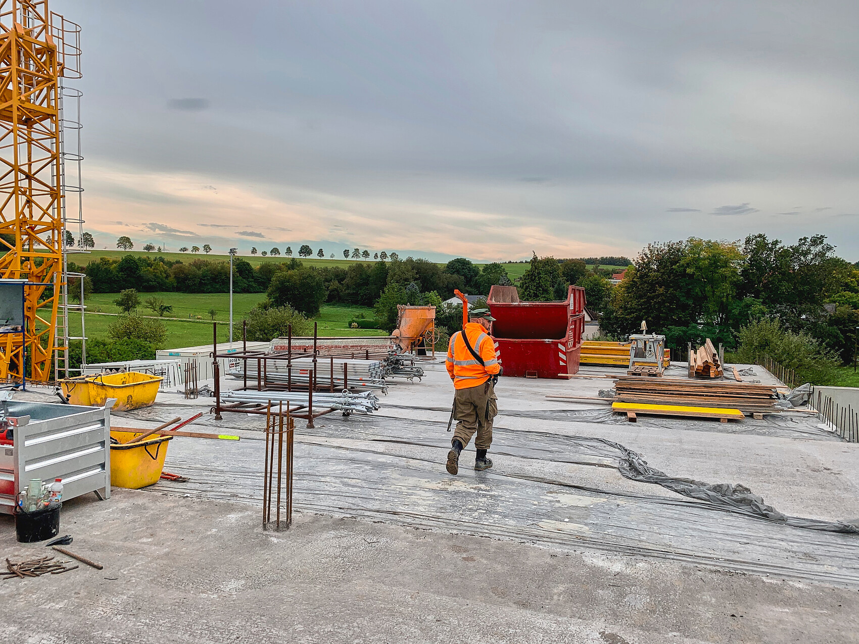 Baustelle: Arbeiter in Warnweste vor Bodenplatte; Bewehrung, Kran links, grüne Landschaft.