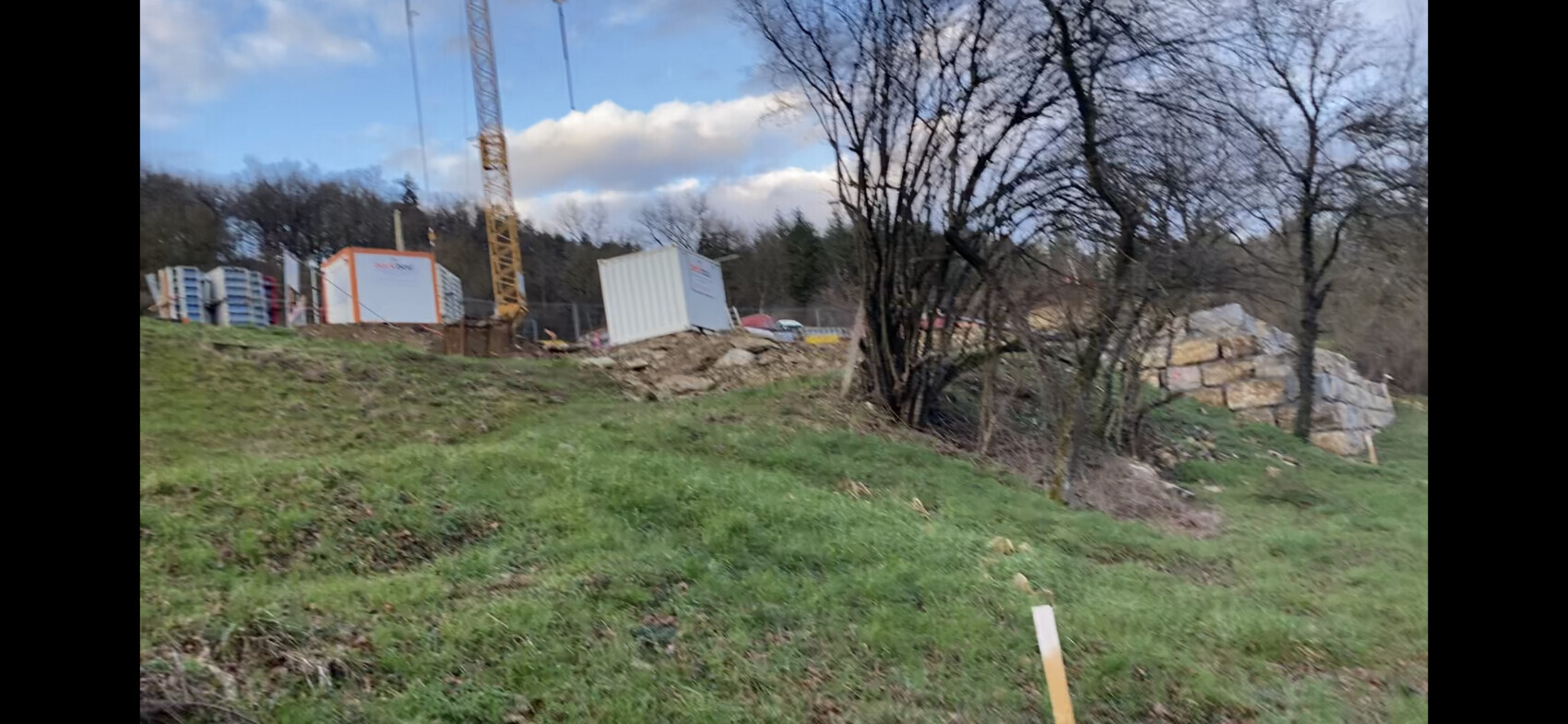 Baustelle mit gelbem Kran, Container und Bauholz auf grasbewachsenem Hang, Bäume im Hintergrund.