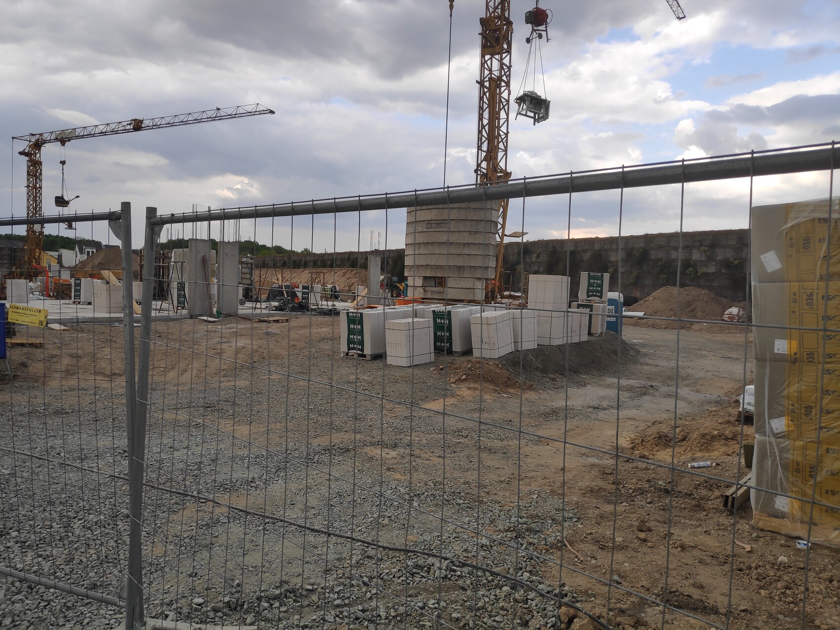 Baustelle mit Kranen, Zaun davor, Betonblöcke und Baumaterialien auf dem Gelände.