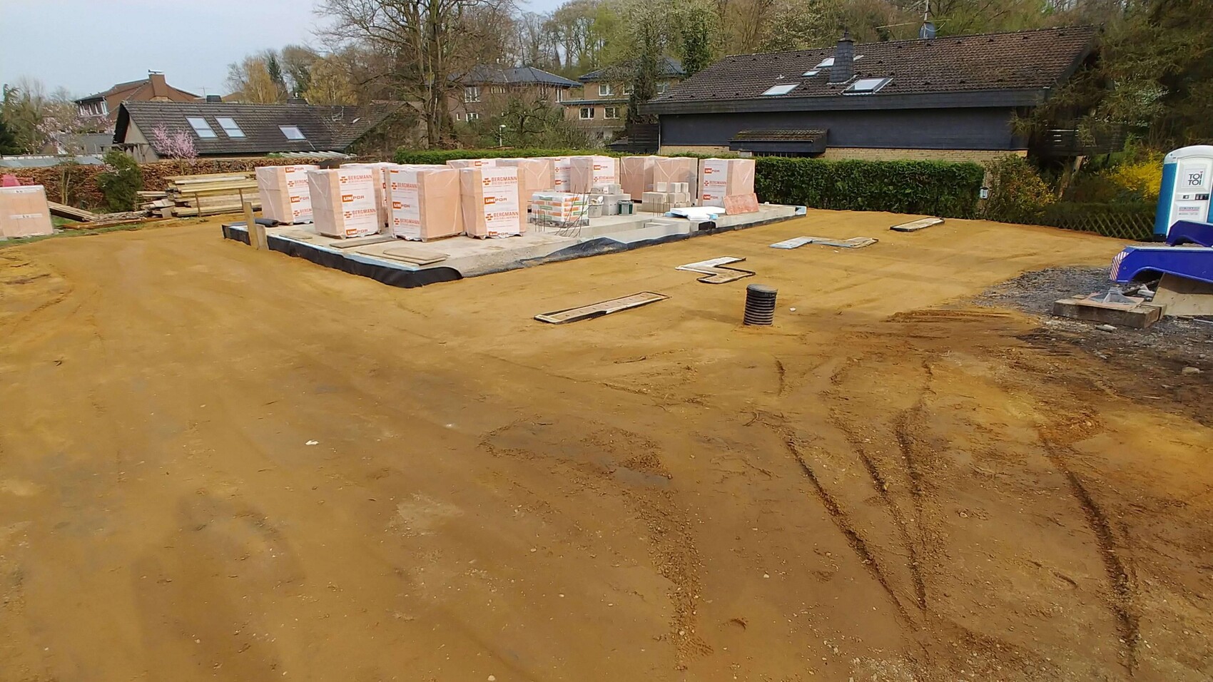 Baustelle mit Erdboden, gestapelte Bauplatten, Palettenmaterialien und Rohrverlegung im Vordergrund.