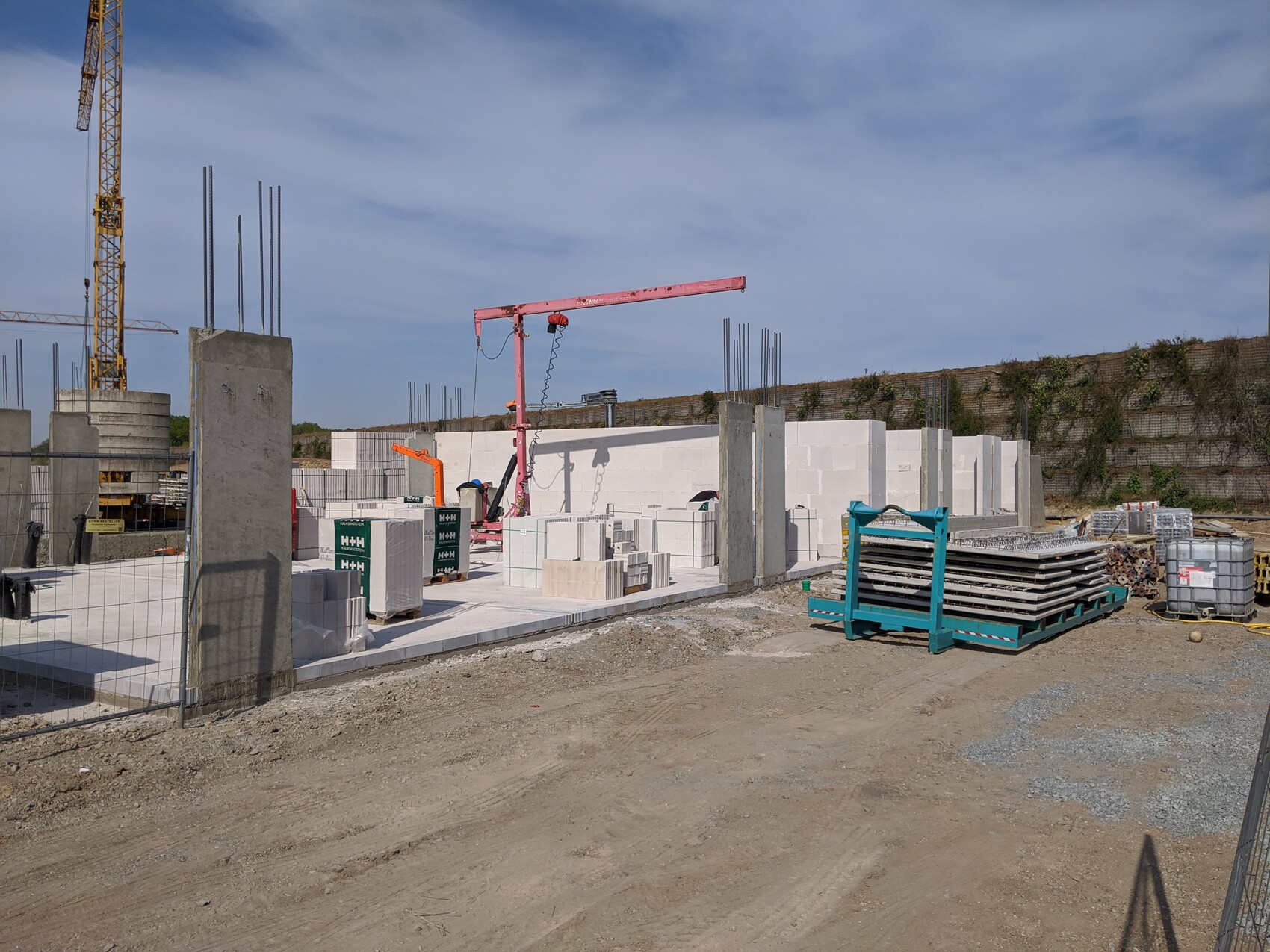 Baustelle mit Betonblöcken, Kran, Stahlstangen und Paletten auf einer offenen Baufläche.