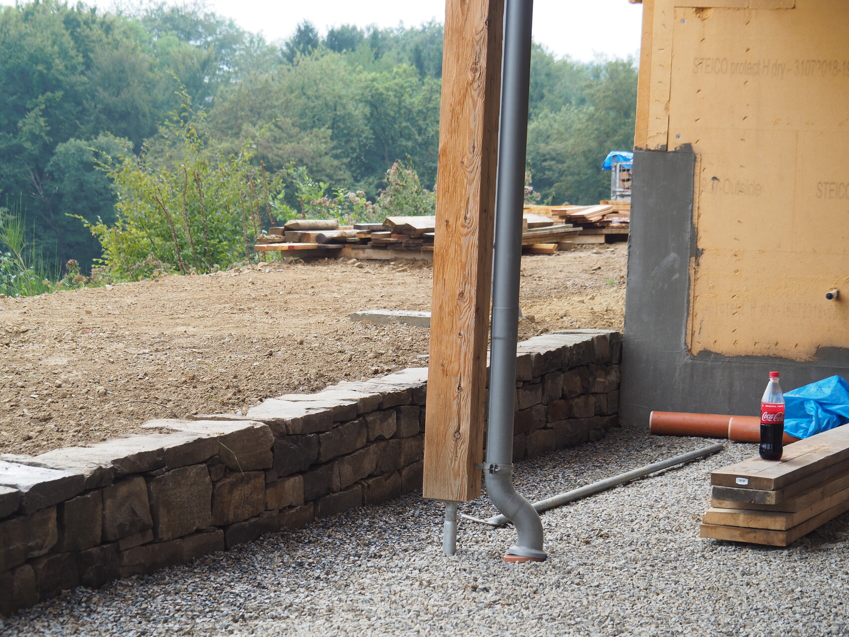 Baustelle mit Steinmauer, Holzpfosten, Fallrohr, Holzstapeln und Cola-Flasche.