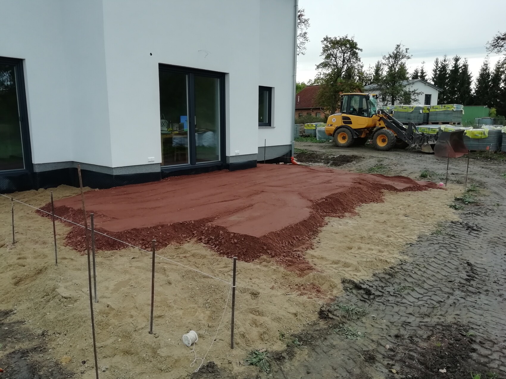 Baustelle am Neubau: roter Erdgrund und Sandfläche, gelber Radlader neben weißem Haus