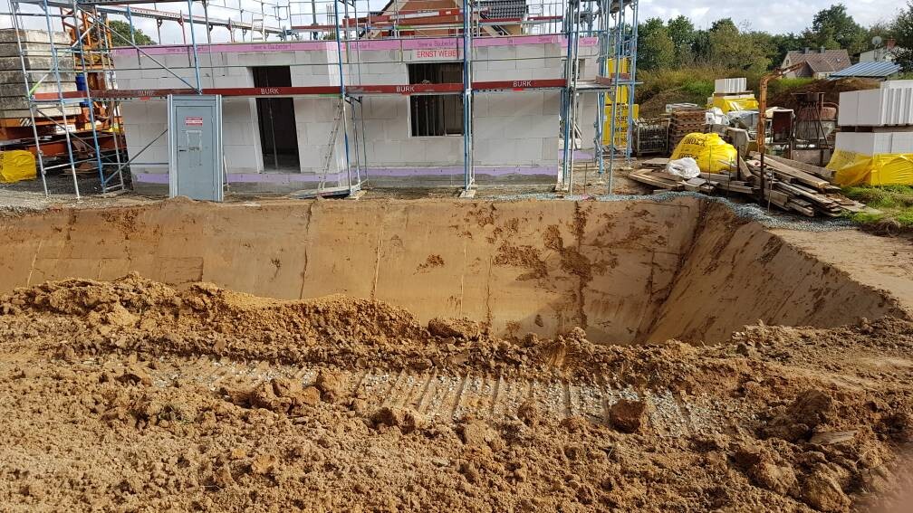 Große Baugrube vor einem neu errichteten Gebäude mit Gerüst und Baumaterialien.