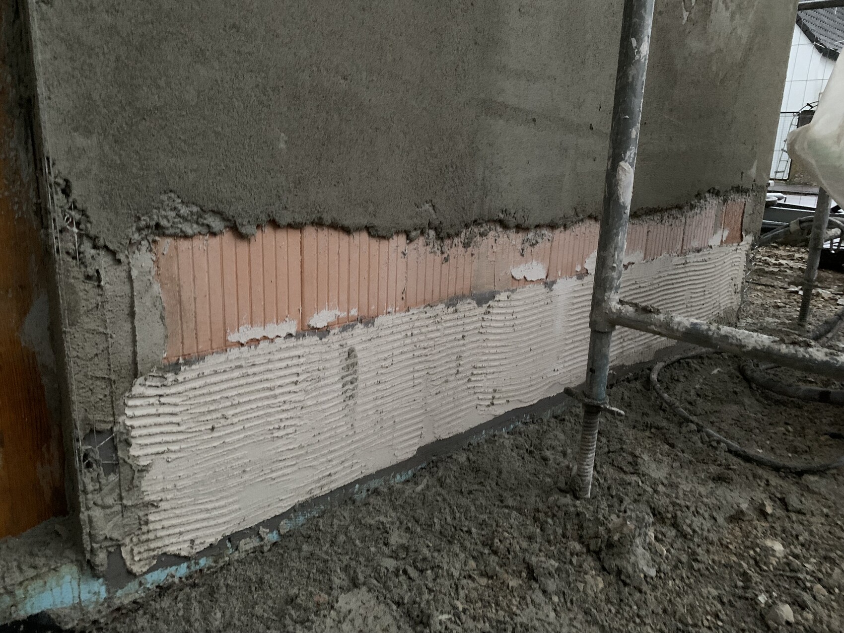 Außenwand-Baustelle: Putzabbruch, rosafarbene gerillte Dämmplatten sichtbar; Gerüststange.