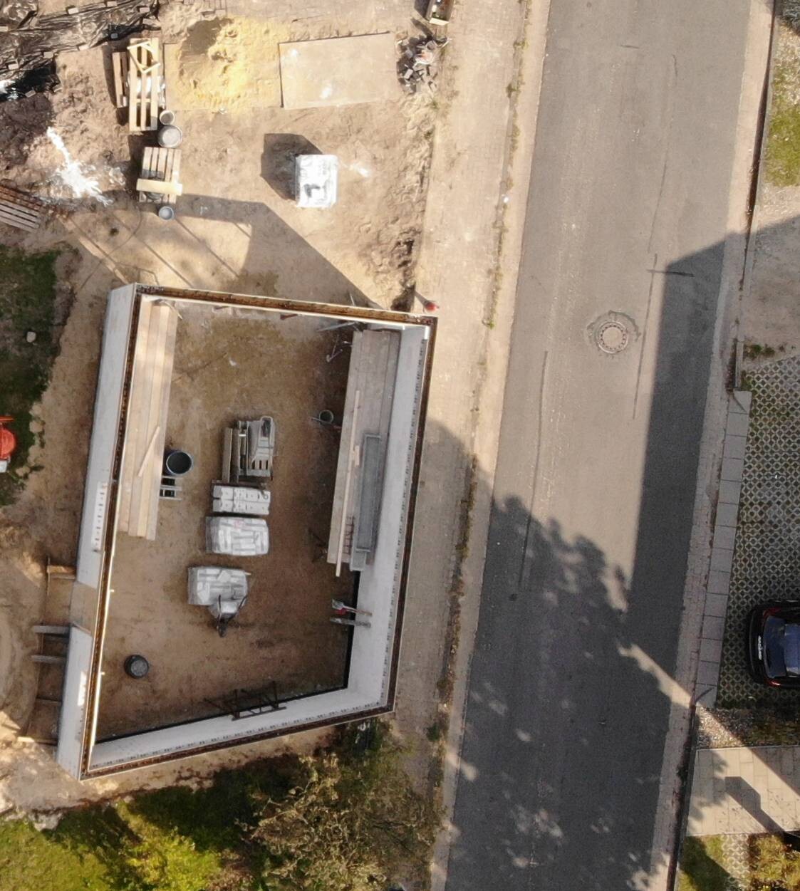 Luftaufnahme eines Bauplatzes mit rechteckigem Rohbau, Baumaterialien und Straße daneben.
