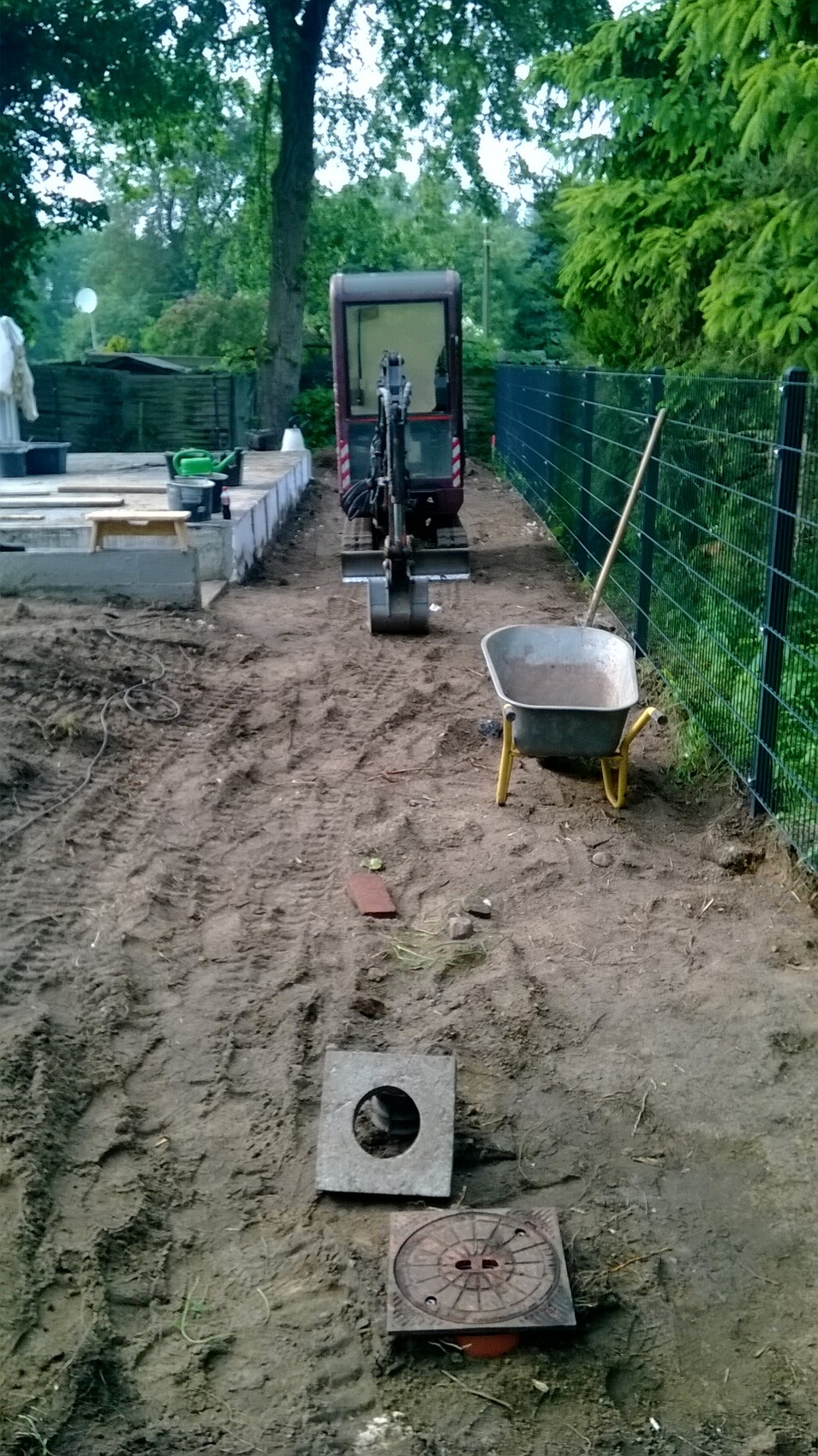 Kleiner Garten-Baustelle mit Mini-Bagger, Schubkarre und offenem Kanaldeckel auf Erdboden.