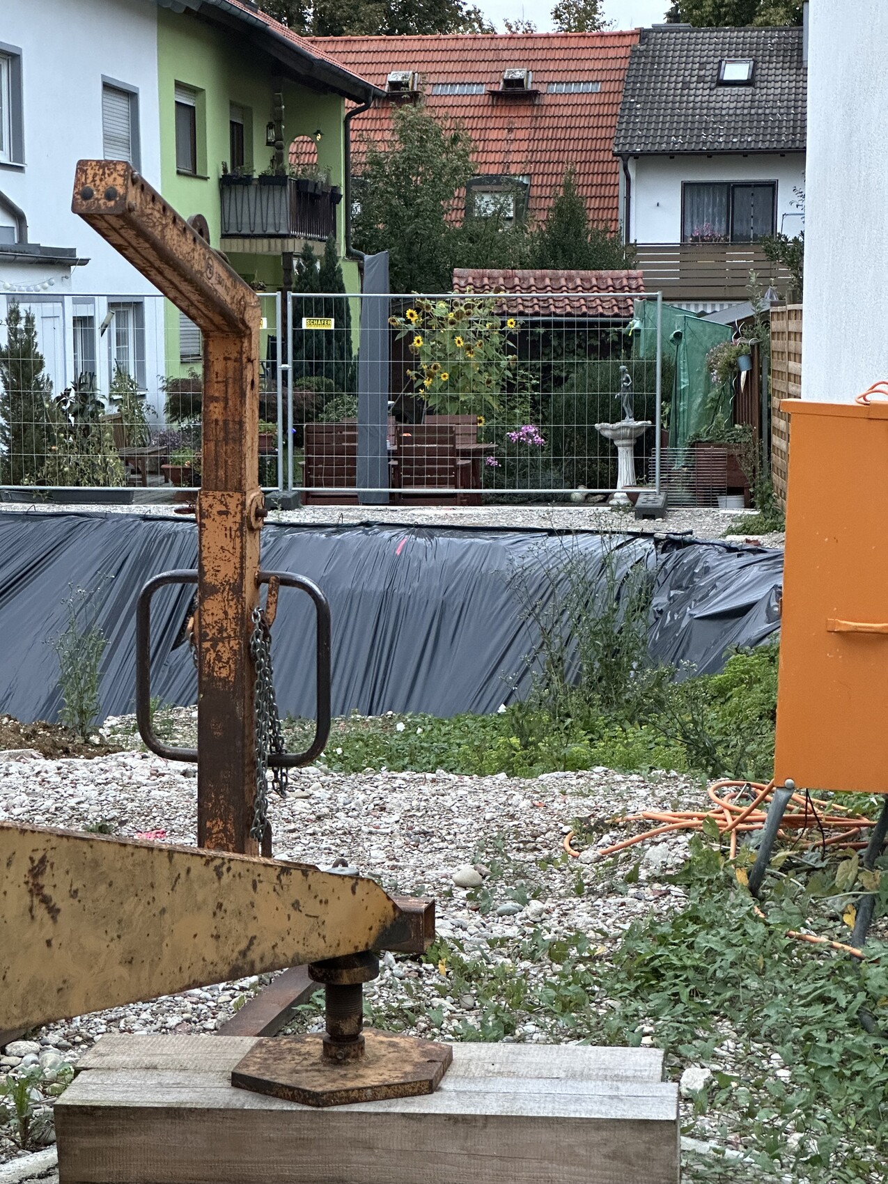 Rostige Baumaschine mit Hebearm im Vordergrund, Baugrube mit schwarzer Folie dahinter, Garten hinter Zaun.