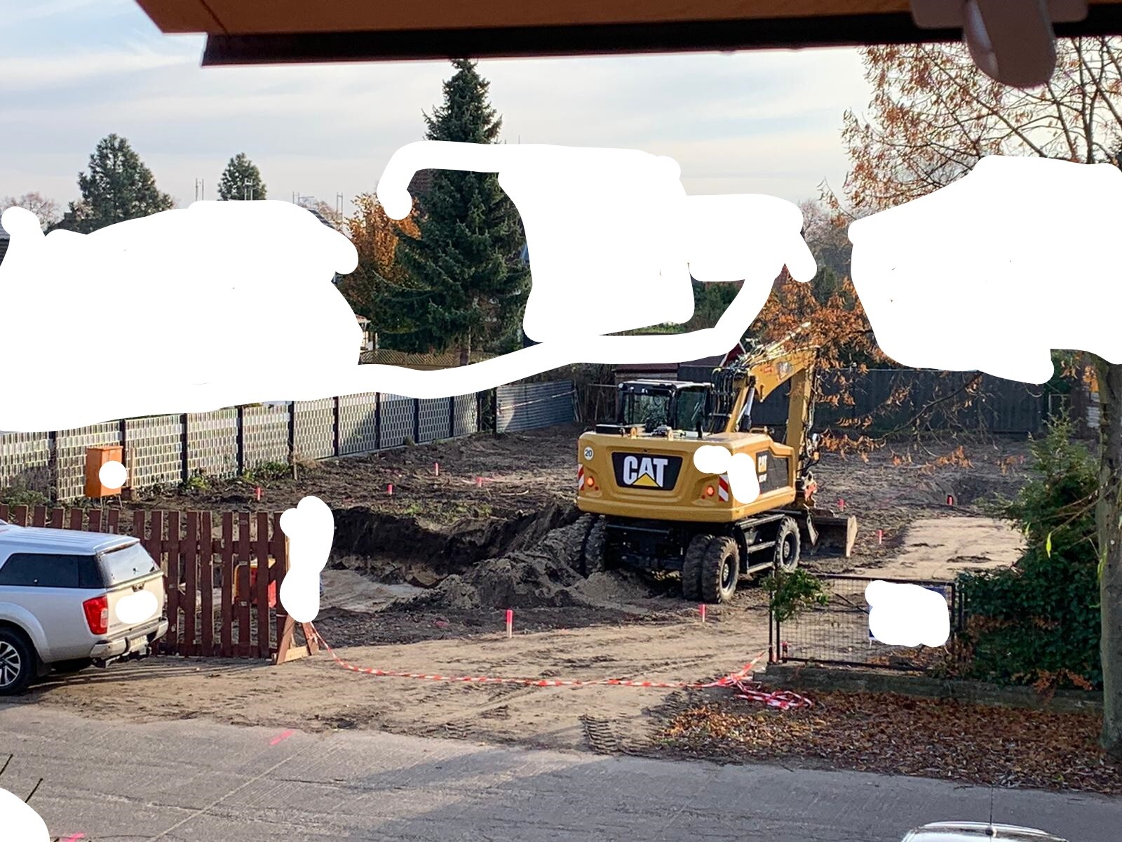 Gelber CAT-Bagger auf Baustelle hinter Zaun, links ein Auto, Absperrbänder markieren das Gelände.