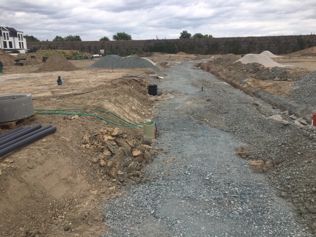 Baustelle mit Kiesweg, Erdhaufen, Rohren und einer Mauer im Hintergrund.