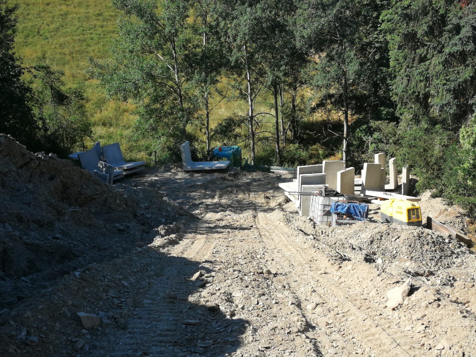 Baustelle im Wald am Schotterweg mit Betonblöcken, Baugeräten und verstreuten Möbeln.