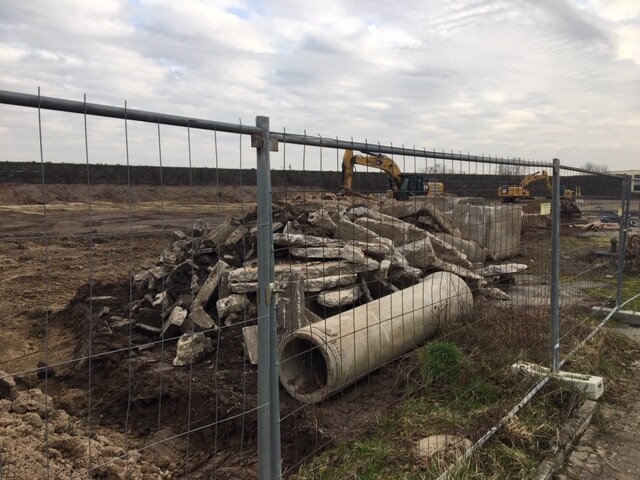 Baustelle mit Betonbruch, einem großen Rohr, Bauzaun im Vordergrund, Bagger im Hintergrund.
