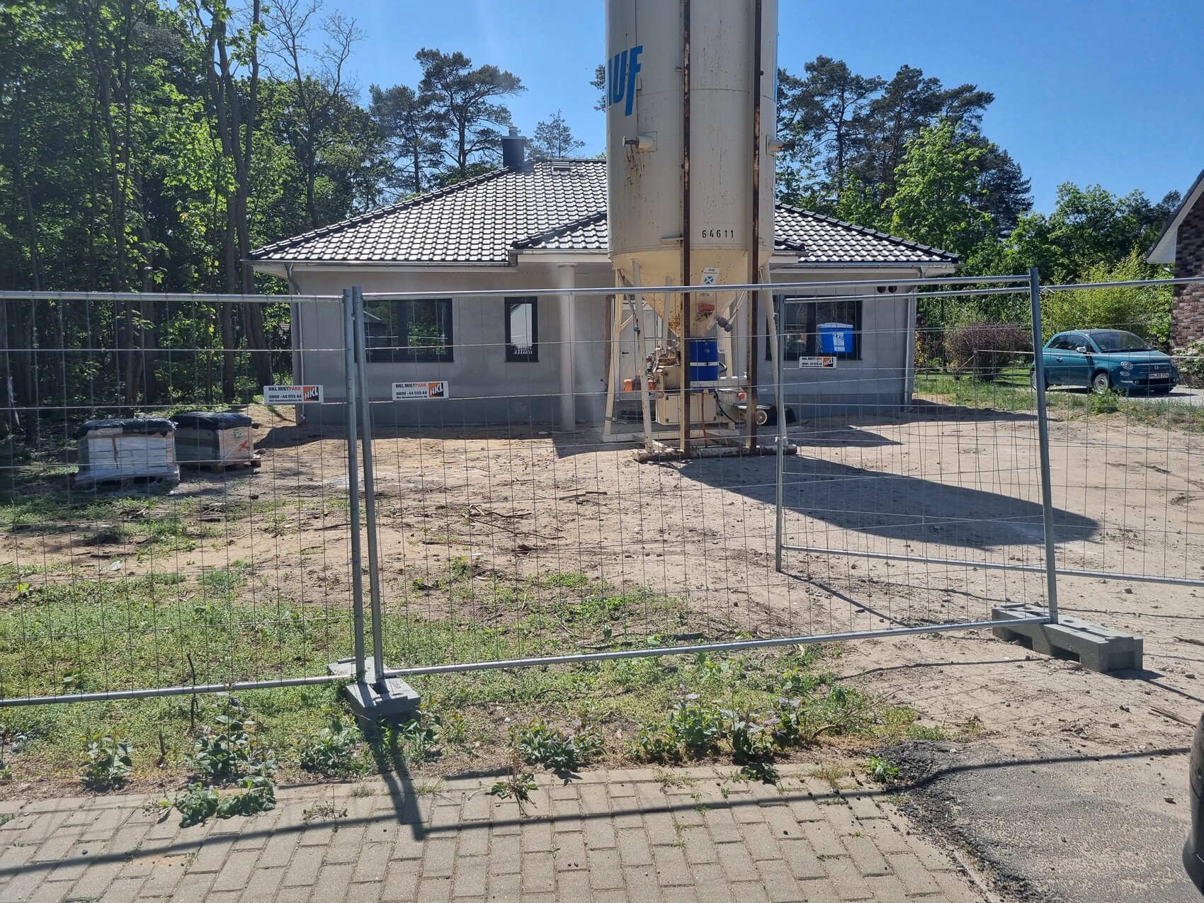 Baugelände hinter Zaun: Bohrturm neben kleinem Wohnhaus, rechts ein blaues Auto.