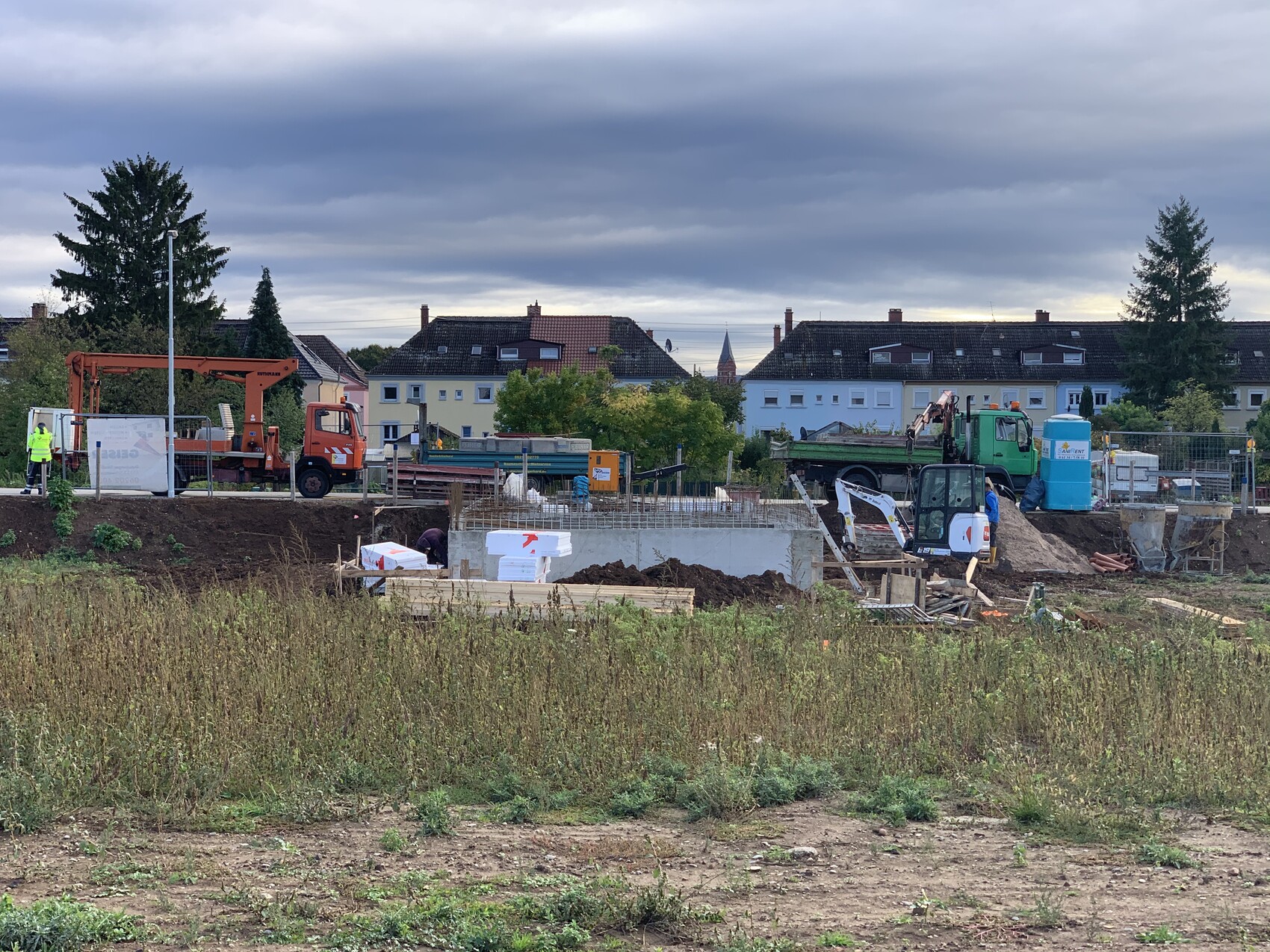 Baustelle mit Bagger, Kran und Baucontainer; Bauarbeiter in Warnkleidung vor Wohnhäusern.