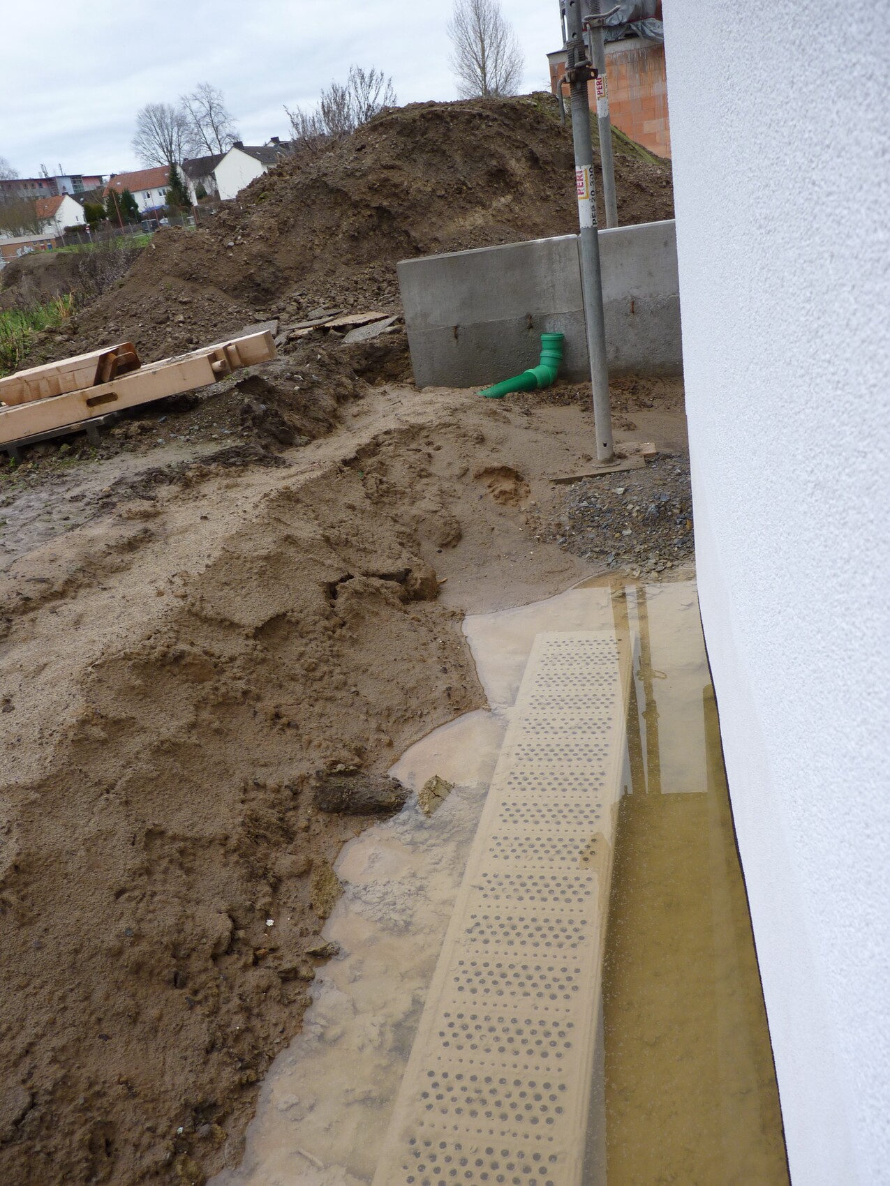 Baustelle mit Erdaushub, grünem Abwasserrohr, perforierter Betonplatte und Pfütze neben Hauswand.