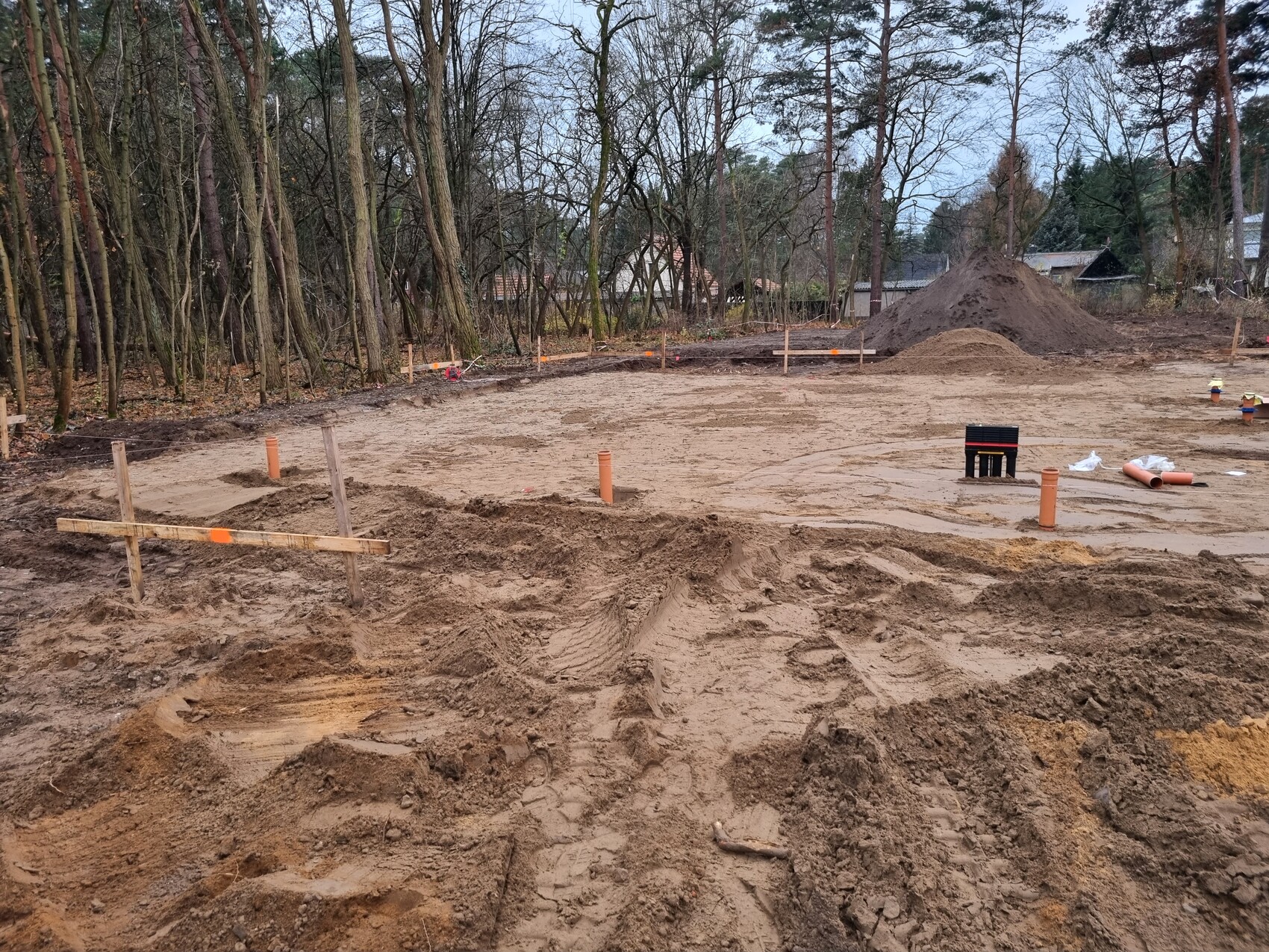 Baustelle im Gelände: Erdarbeiten, Rohre, Holzmarkierungen, Sandhügel, Wald im Hintergrund.