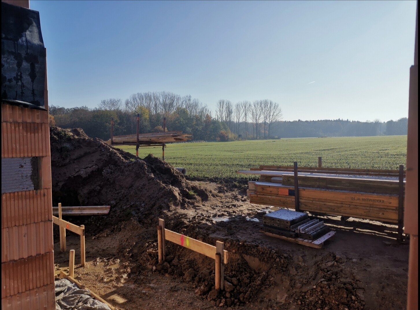 Baustelle im Freien: Erdmaterial, Holzbalkenstapel, linke Backsteinwand, Feldblick
