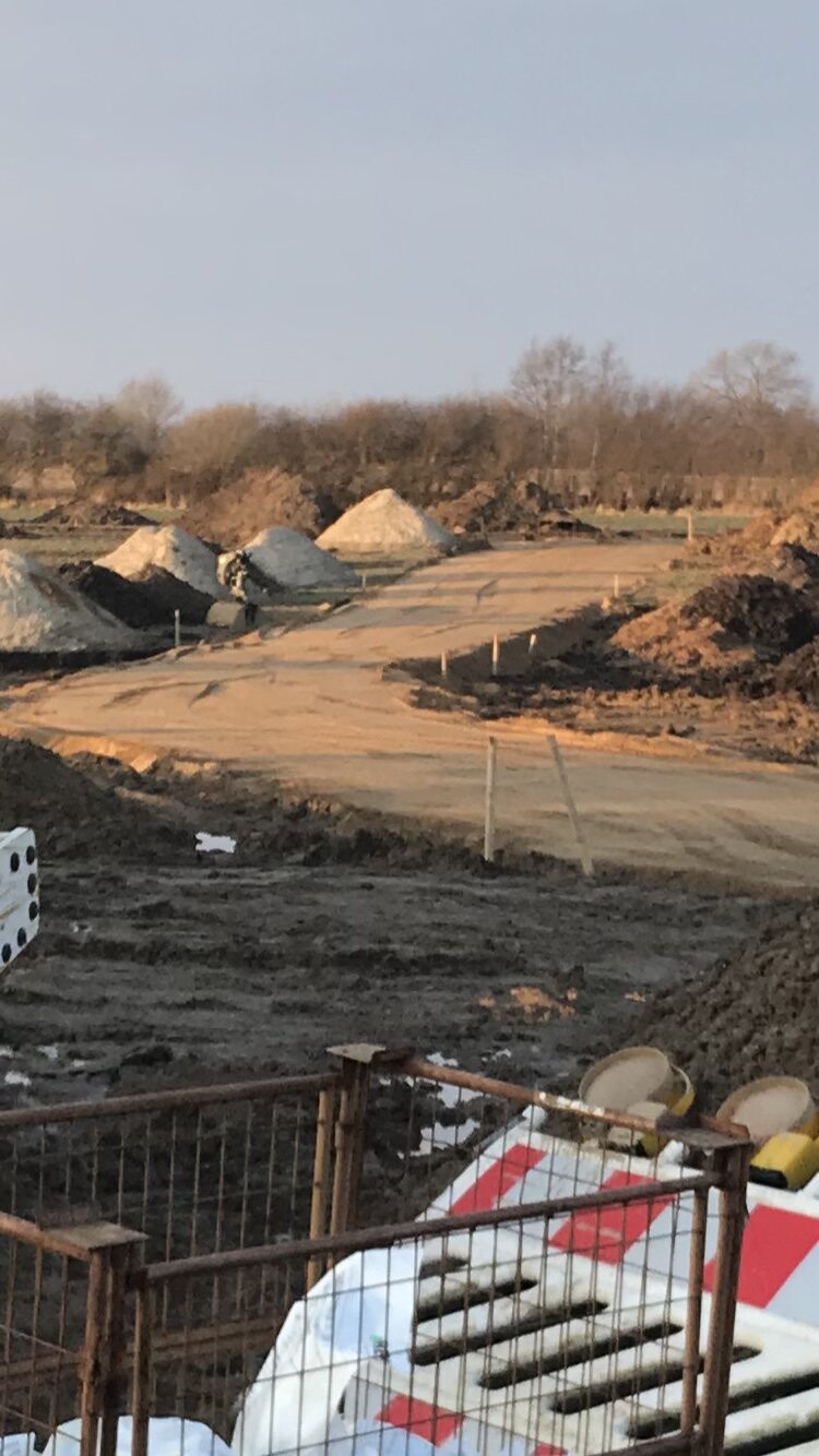 Baustelle mit Sand- und Erdhaufen, unbefestigte Wege, Metallzaun im Vordergrund, Bäume im Hintergrund